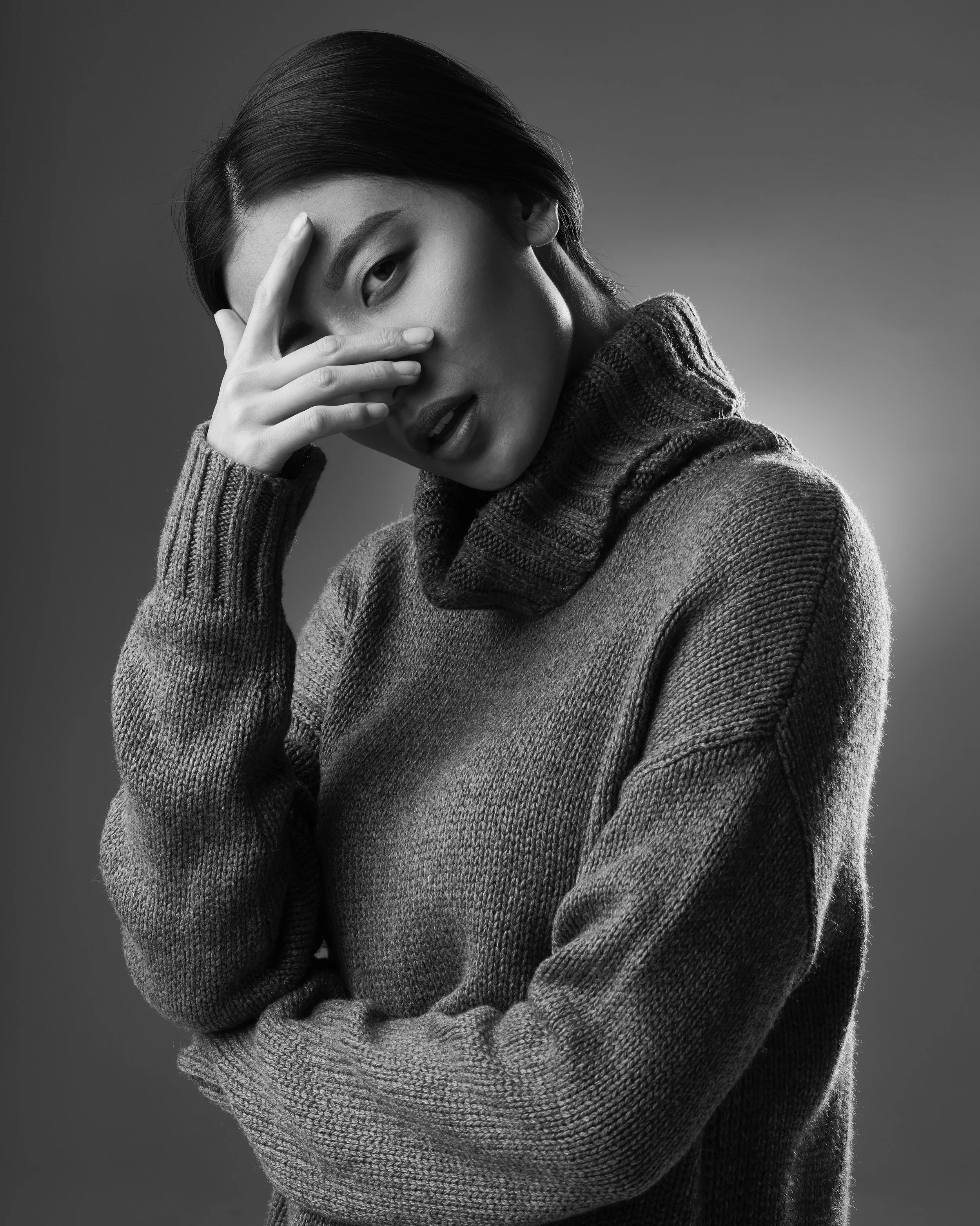 Black and white photo of a young woman with dark hair wearing a textured sweater and turtleneck, making a gesture with her hand near her face.