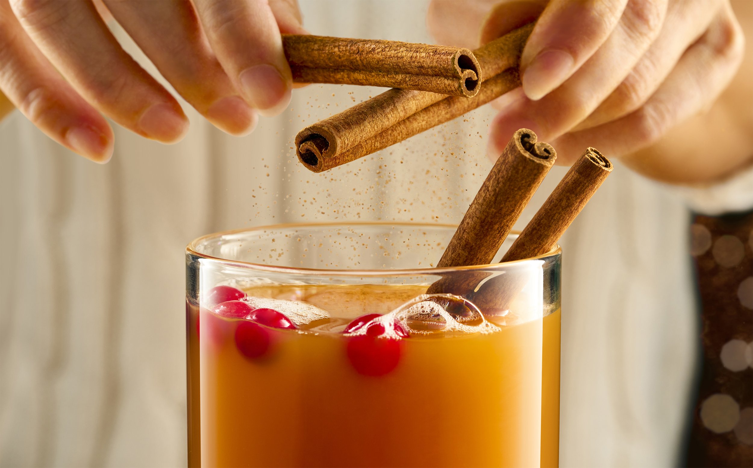Hands sprinkling cinnamon over a glass of orange-colored beverage with red berries, with cinnamon sticks inside the glass.