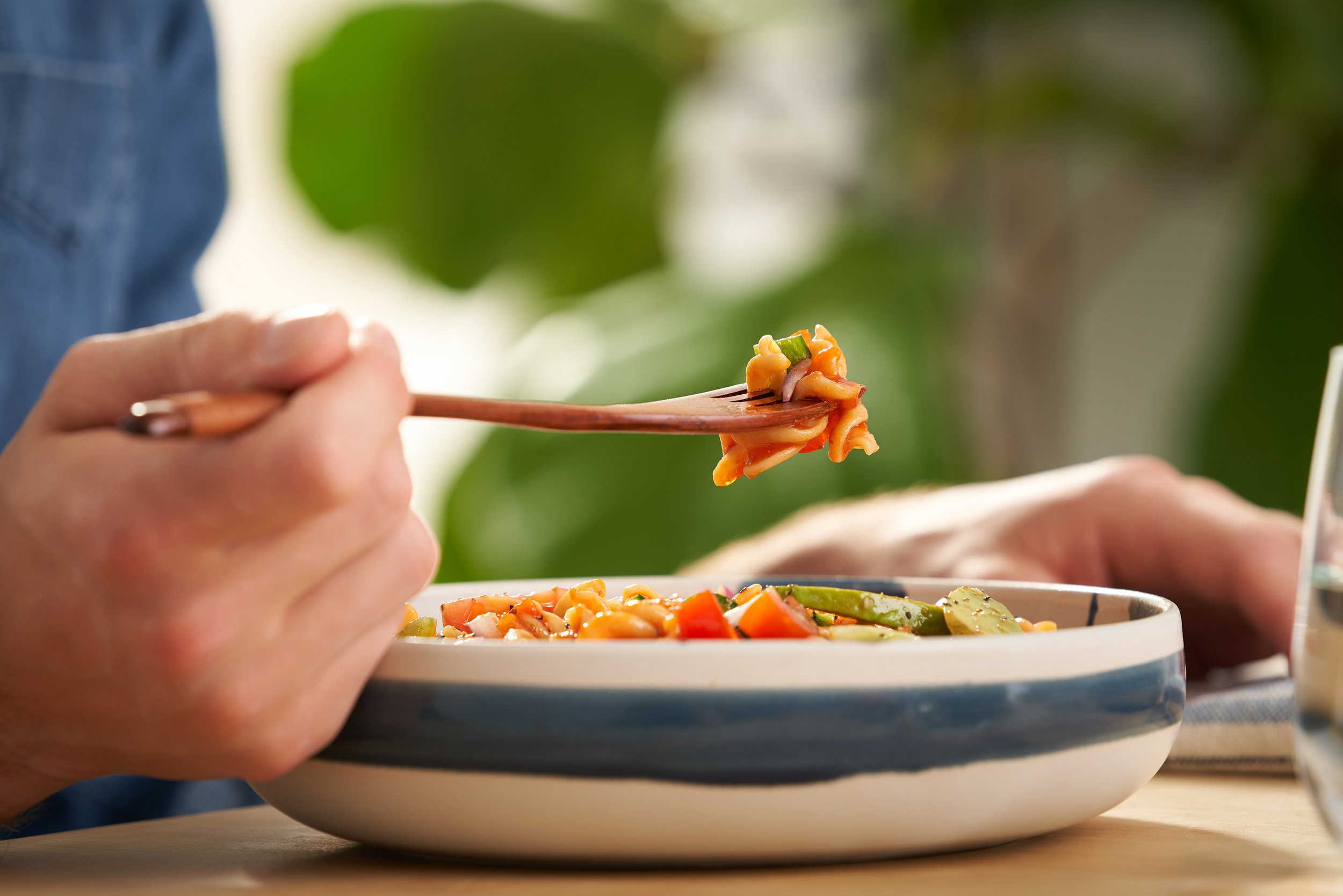 Person holding chopsticks with noodles and vegetables on a ceramic plate, with a blurred background of green foliage.