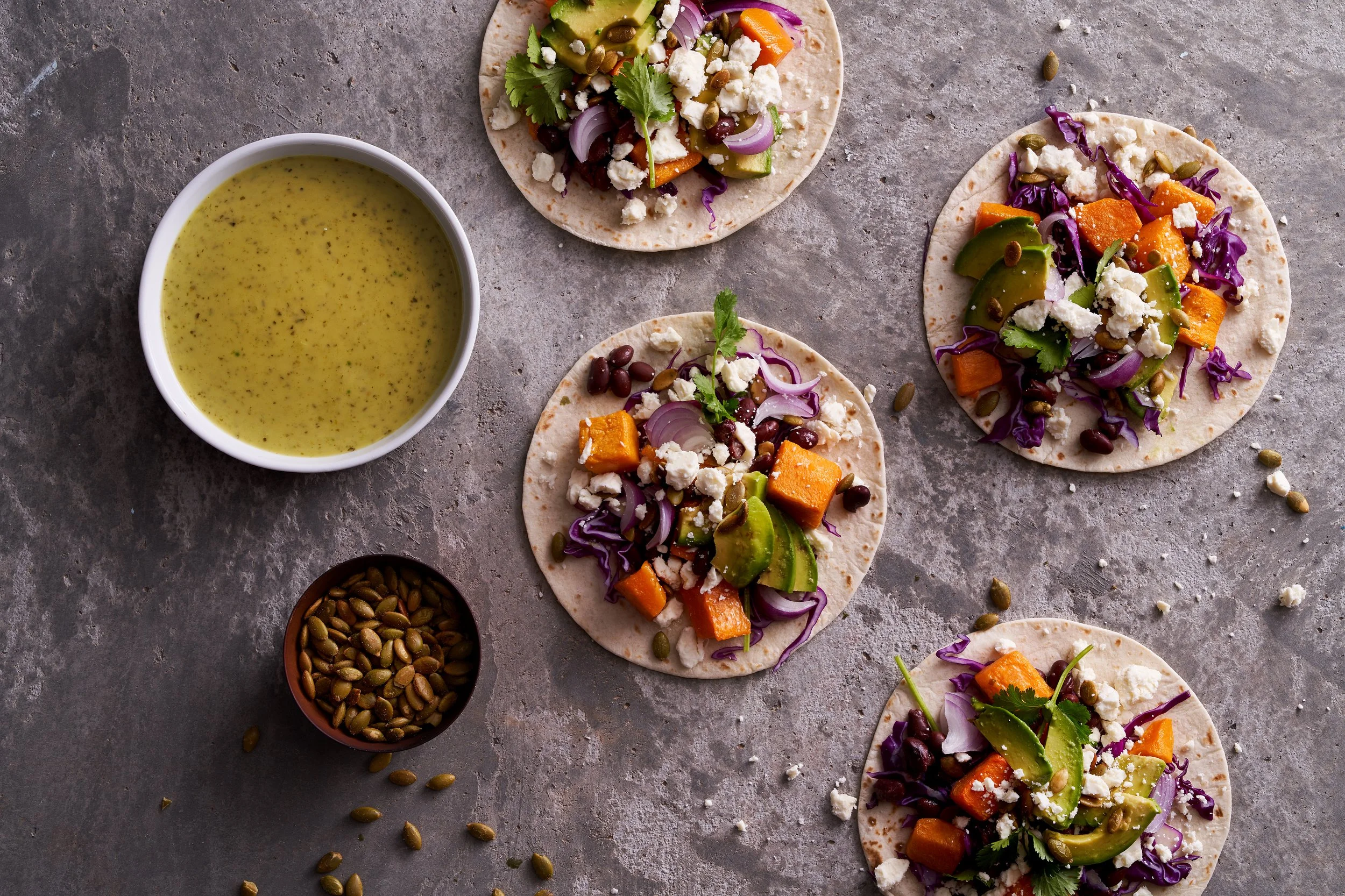 Four small flour tortillas topped with roasted sweet potatoes, avocado slices, red onion, purple cabbage, cilantro, pumpkin seeds, and crumbled cheese, arranged on a gray surface with a bowl of green sauce and a small bowl of pumpkin seeds.