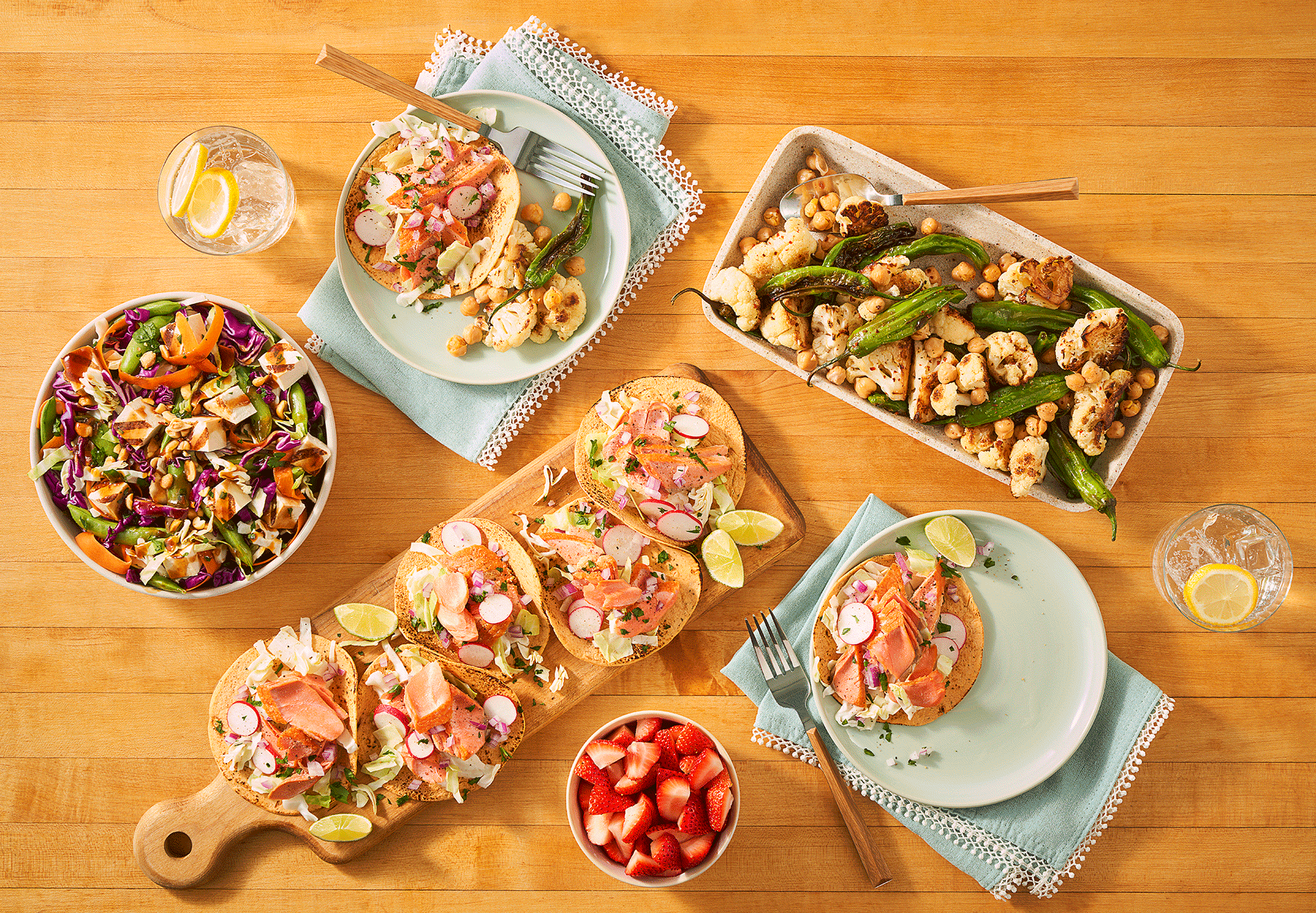Various colorful salads and appetizers on a wooden table, including chopped vegetables, hummus or dip, strawberries, and lemon water.