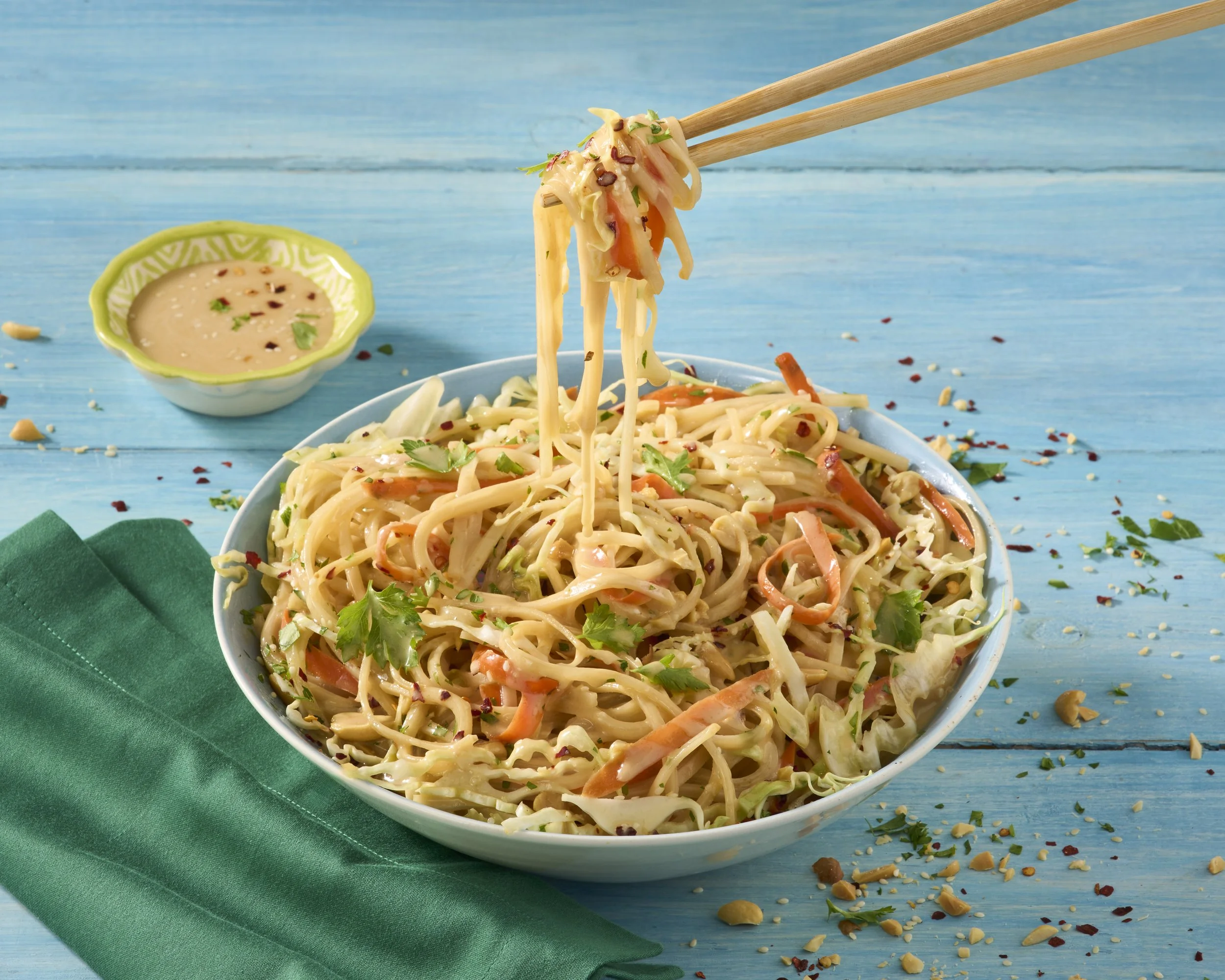 A bowl of Chinese-style cold sesame noodle salad with chopped vegetables and cilantro on a blue wooden surface, with chopsticks lifting noodles and a small bowl of sesame dressing on the side.