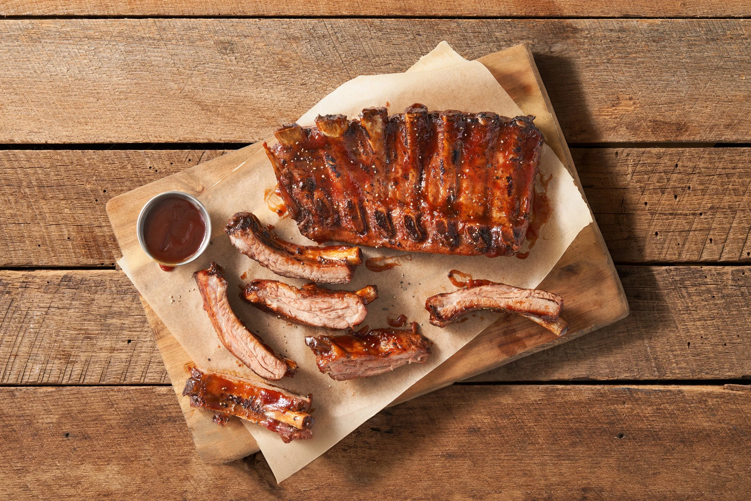 Barbecue ribs served on a wooden table with a small container of barbecue sauce.