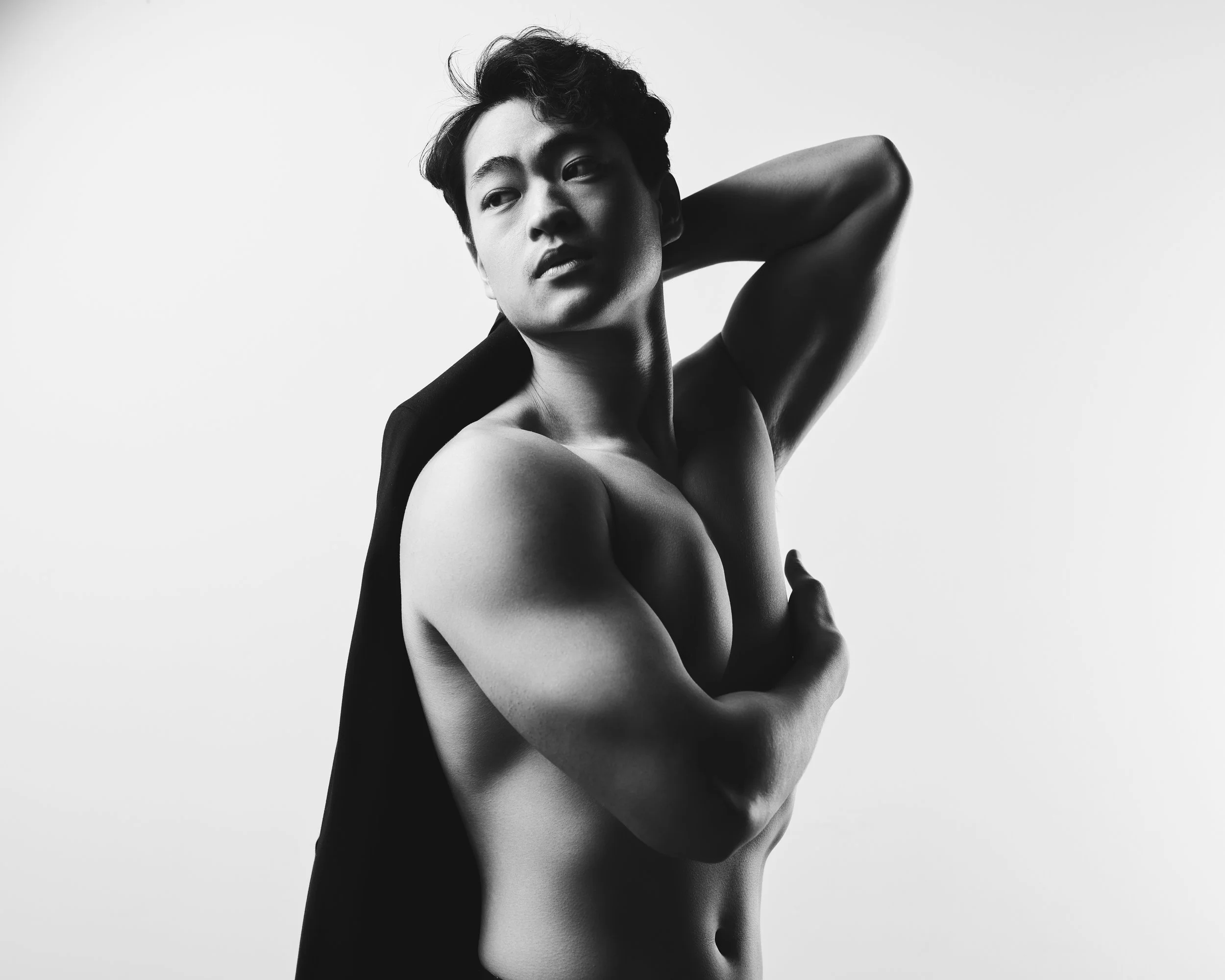 Black and white portrait of a shirtless young man with styled hair, posing with one arm behind his head and the other arm crossed over his chest.