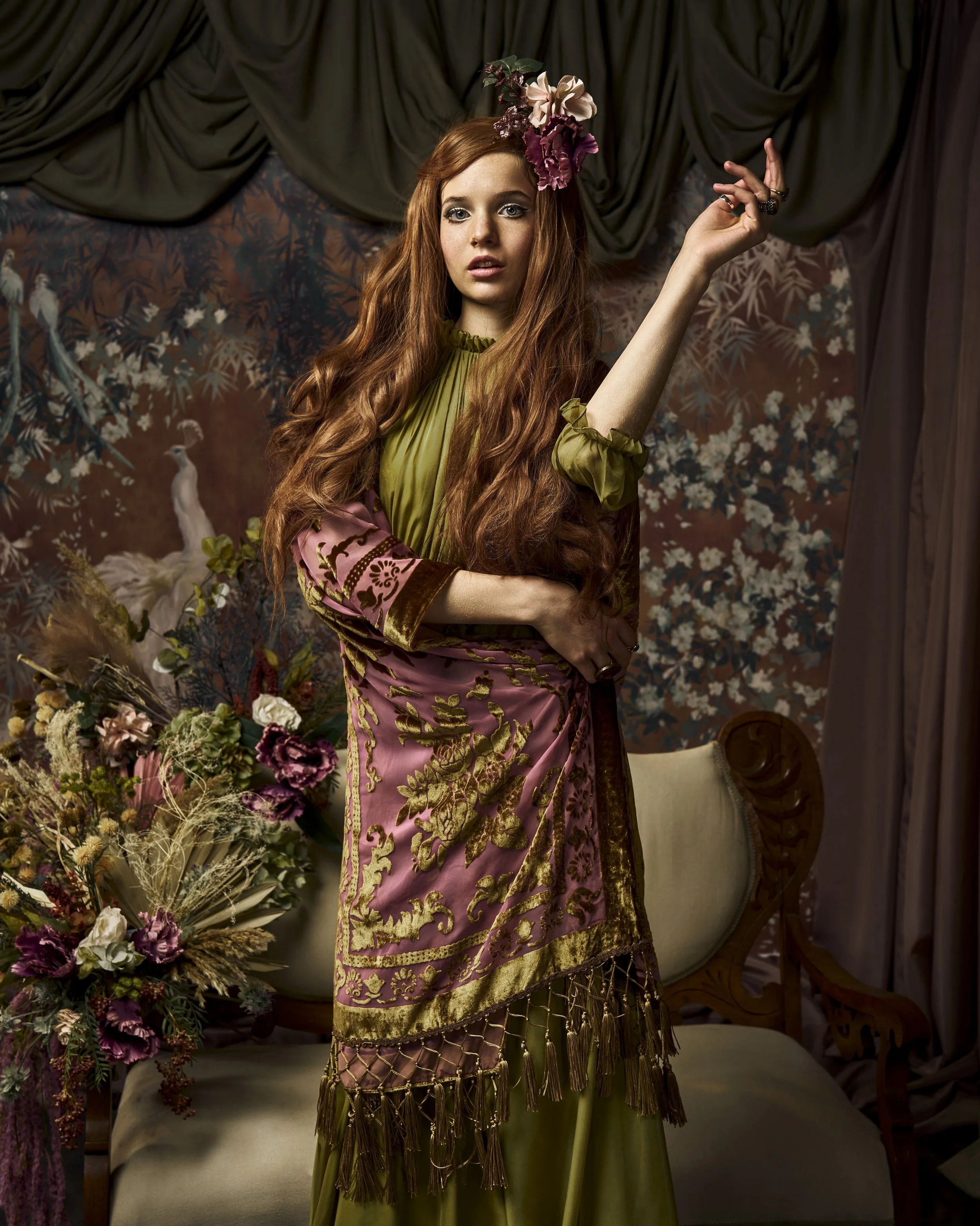 A woman with long curly red hair, wearing a floral headpiece and an elaborate vintage-style dress, stands in a richly decorated room with dark curtains, floral arrangements, and an ornate backdrop featuring a peacock painting.
