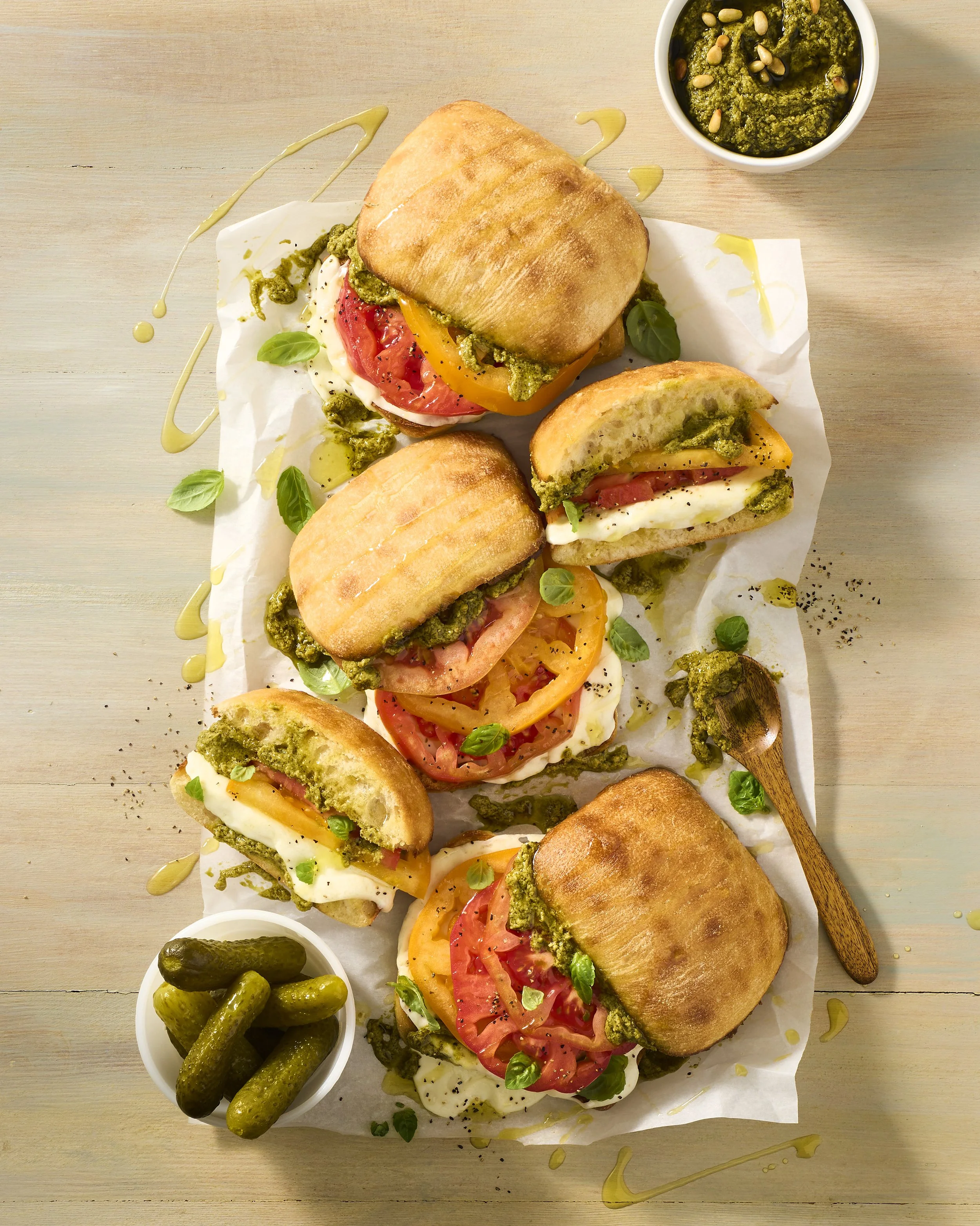 A platter with five sandwiches filled with tomatoes, eggs, and green pesto sauce, garnished with basil leaves, with a side of pickles, served with green pesto sauce.