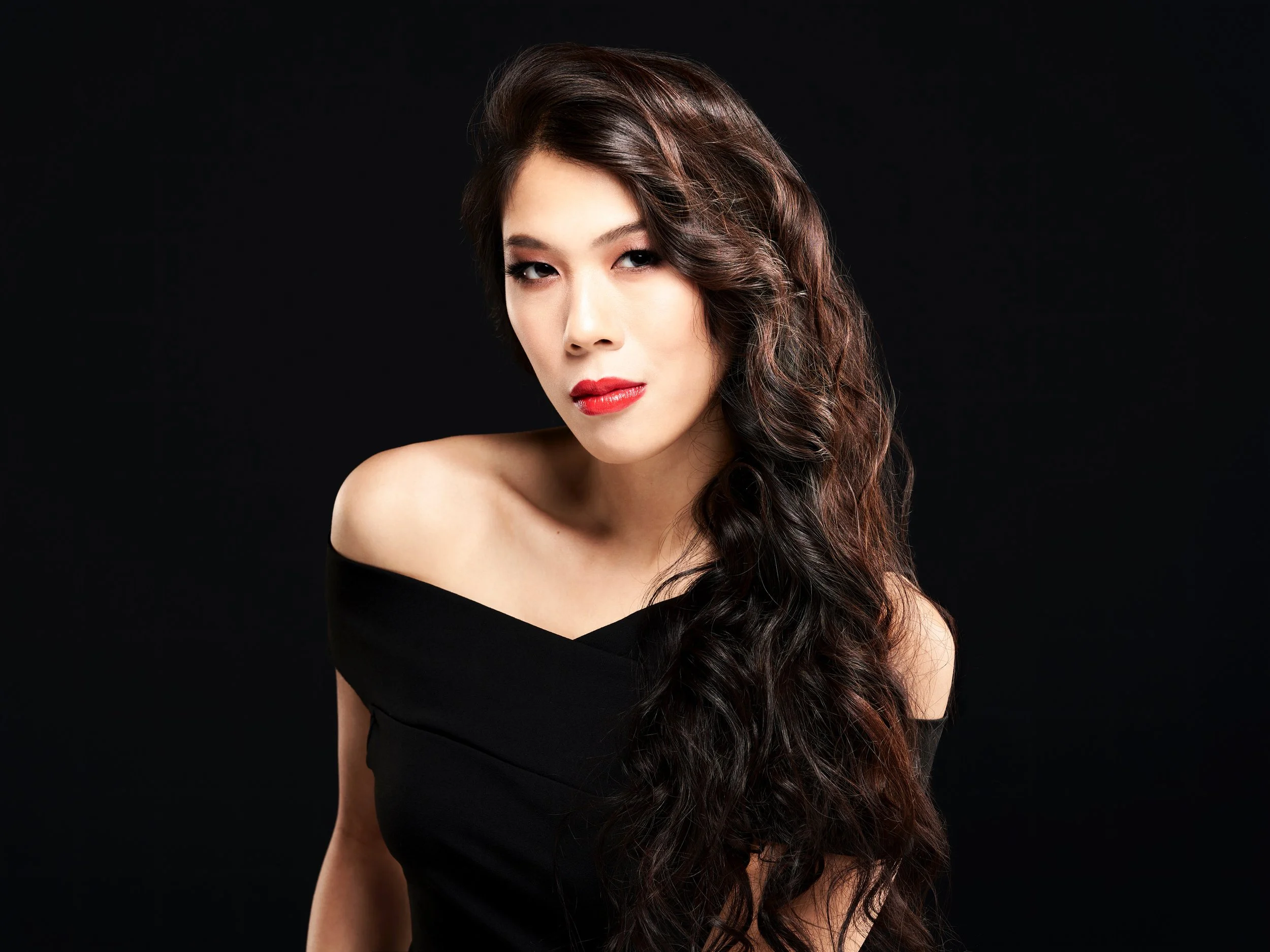 A woman with long wavy brown hair and makeup, wearing a black off-the-shoulder top, poses against a black background.