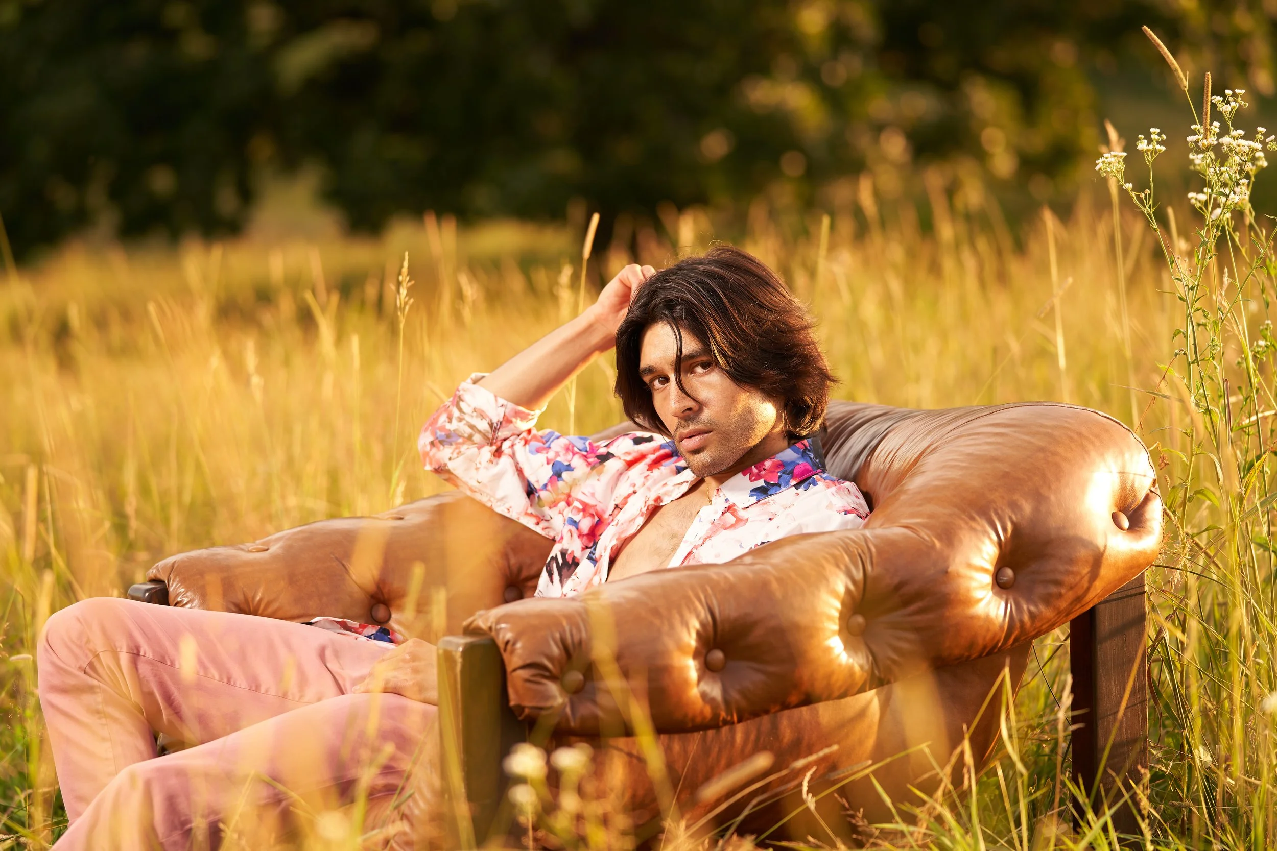 A man with long dark hair reclining on a vintage brown leather sofa in a field of tall grass during sunset.