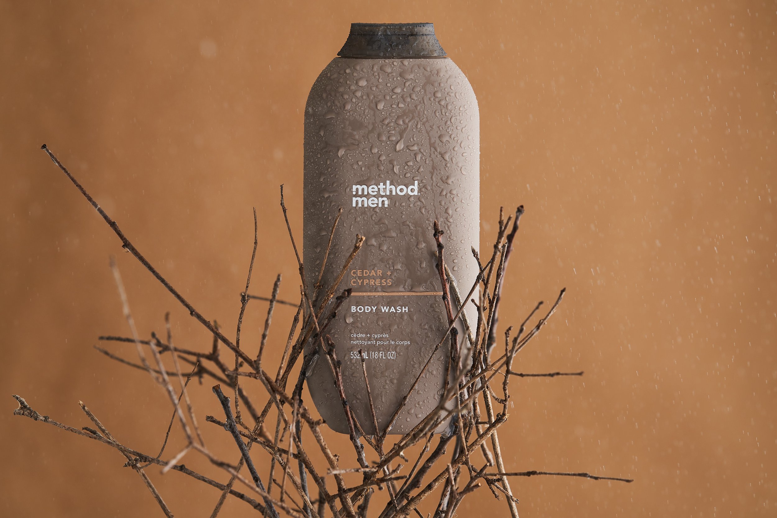 A bottle of Method Men Cedar + Cypress Body Wash surrounded by dry twigs, with water droplets on the bottle, on a brown background.