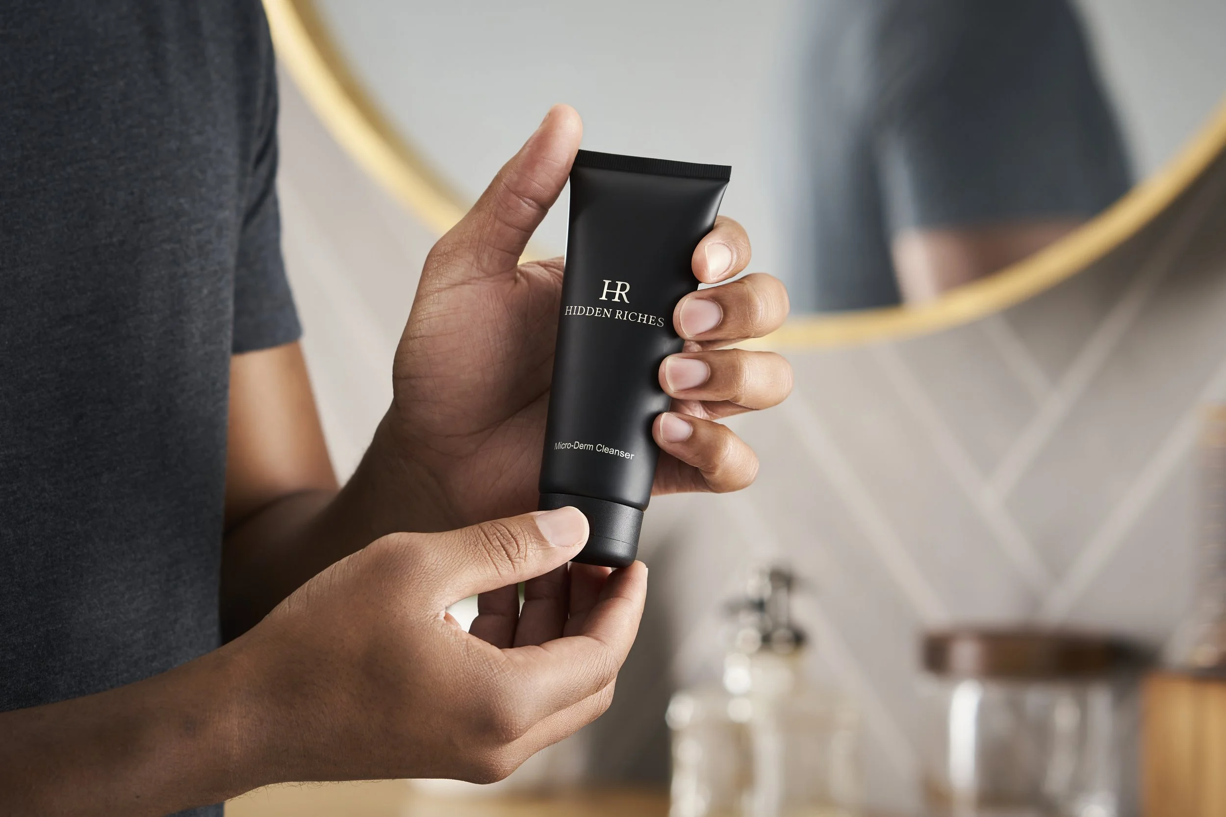 Person holding a black tube labeled 'HR Hidden Riches Micro-Derm Cleanser' in a bathroom with a mirror and bathroom items in the background.