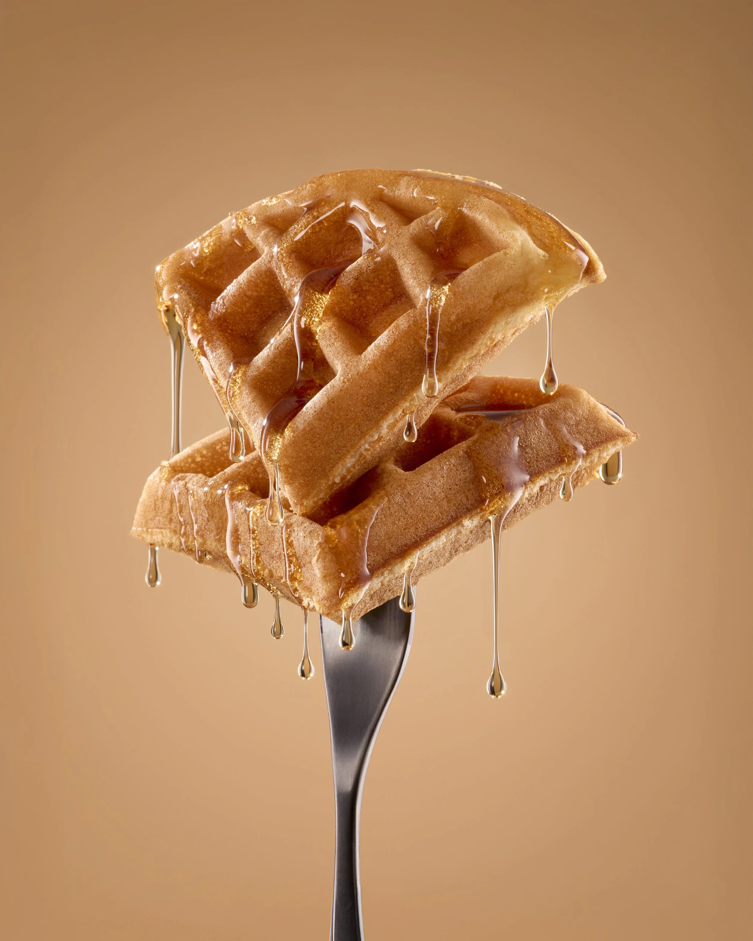 Close-up of two golden brown waffles with syrup, on a fork, dripping with syrup, against a tan background.