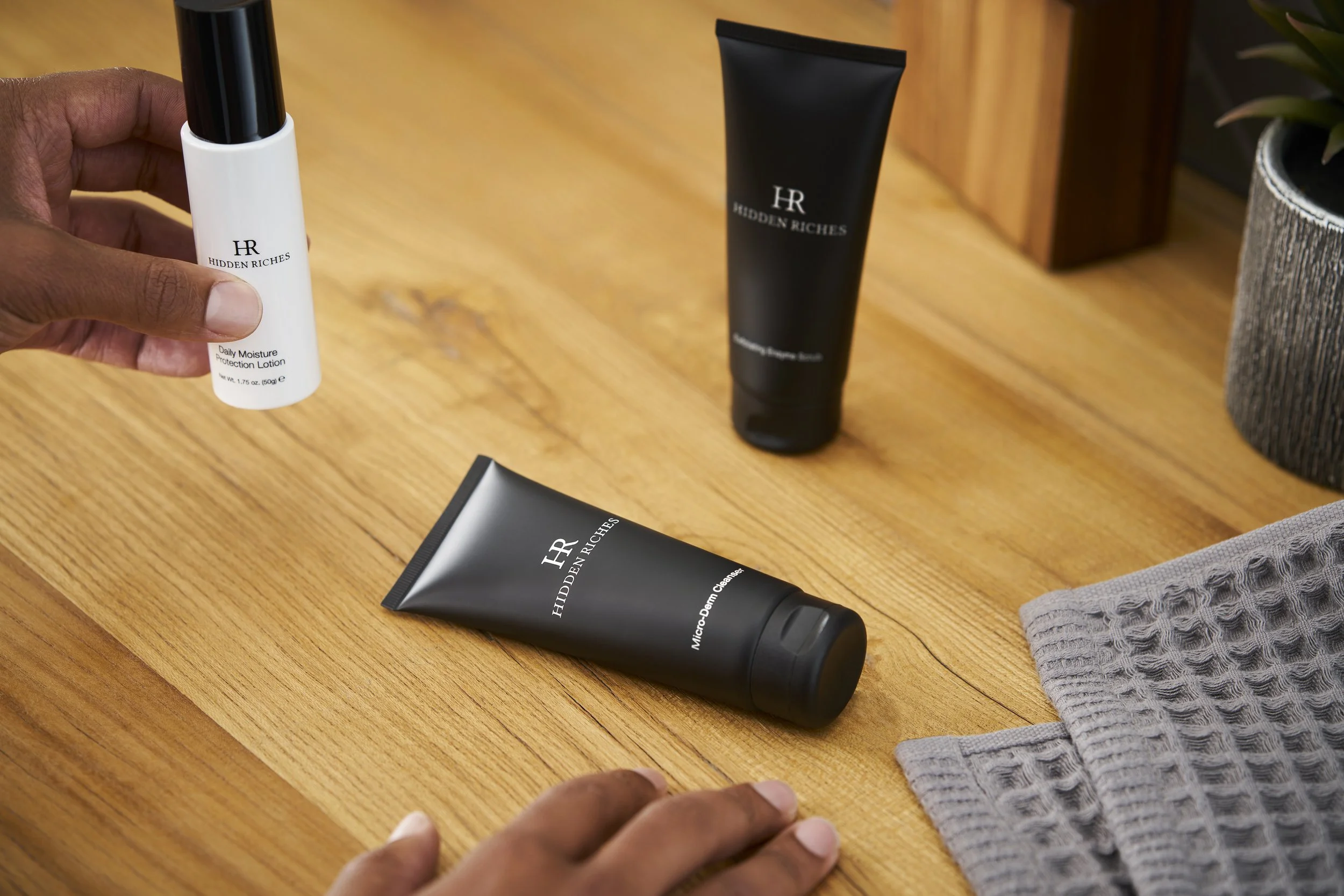 Person holding a bottle of Daily Moisture Protection Lotion from Hidden Riches on a wooden surface along with other skincare products, including a tube labeled Micro-Derm Cleanser, a black tube, and a cloth.