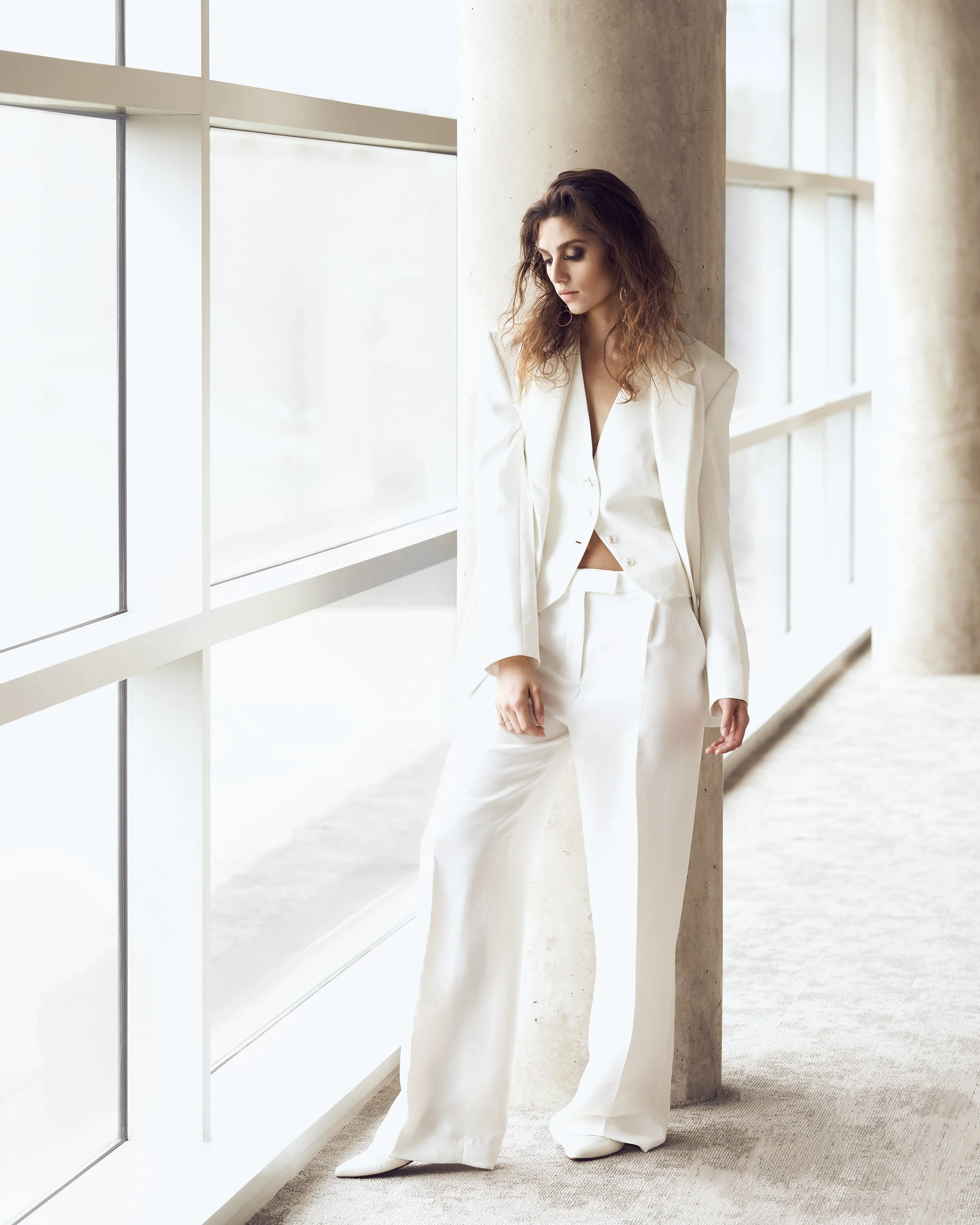 Woman in a white blazer and matching pants standing near a large window with a concrete column in a bright, modern building.
