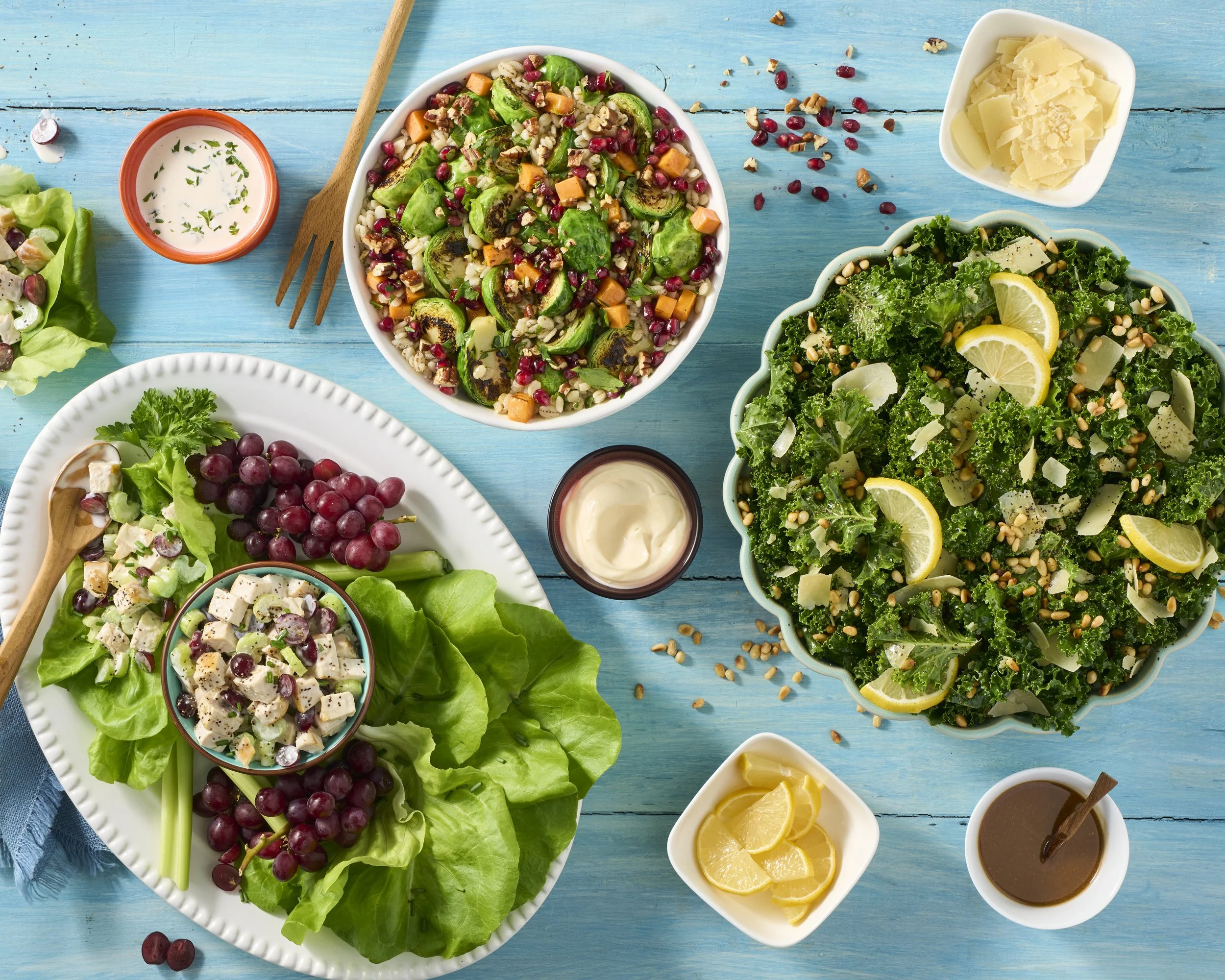 Assorted fresh salads on blue wooden table, including kale salad with lemon slices, mixed grain salad with pumpkin seeds, and fruit and chicken salad, with small bowls of dressing and lemon slices.
