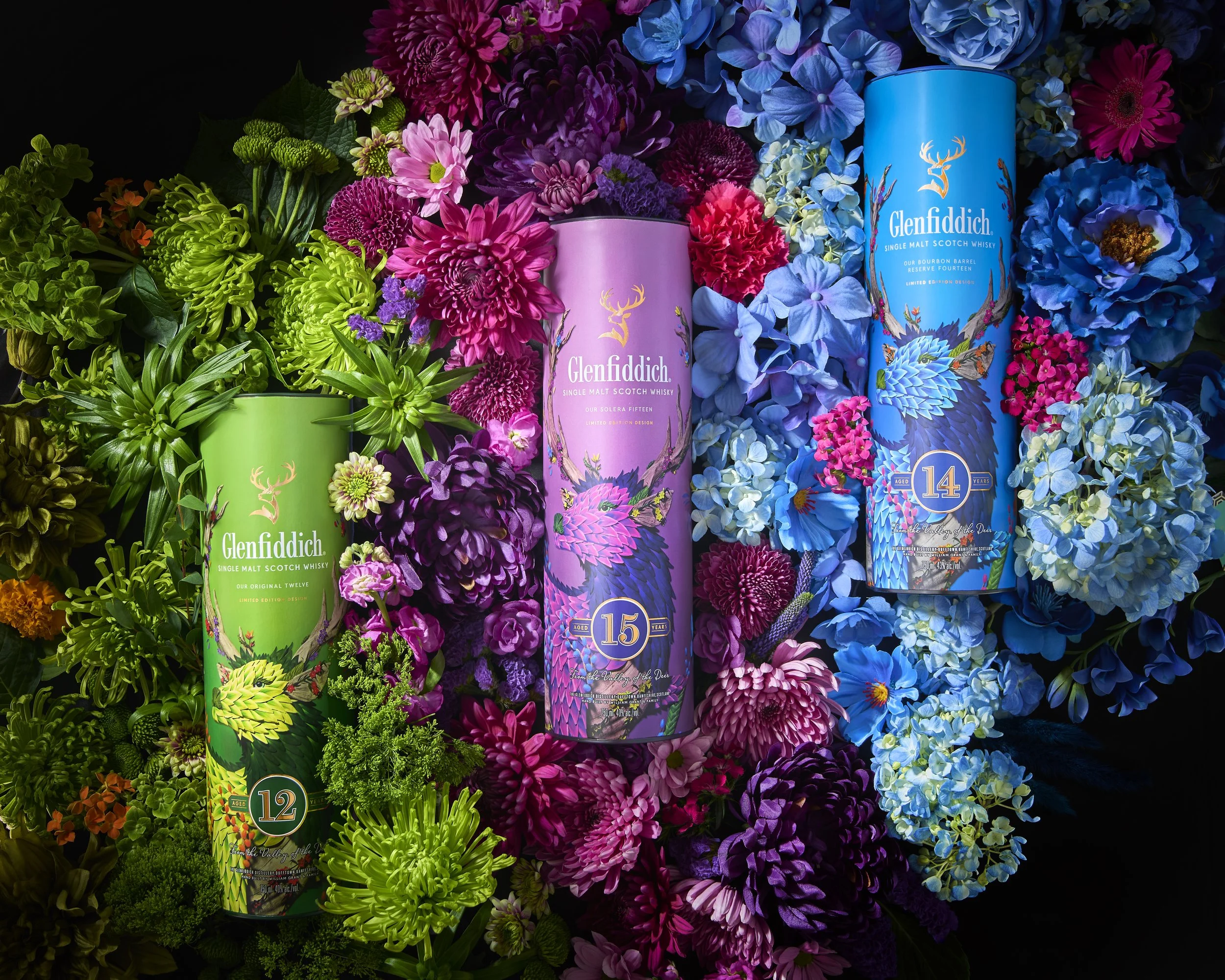 Three colorful Glenfiddich whisky bottles surrounded by vibrant flowers in green, pink, purple, and blue.