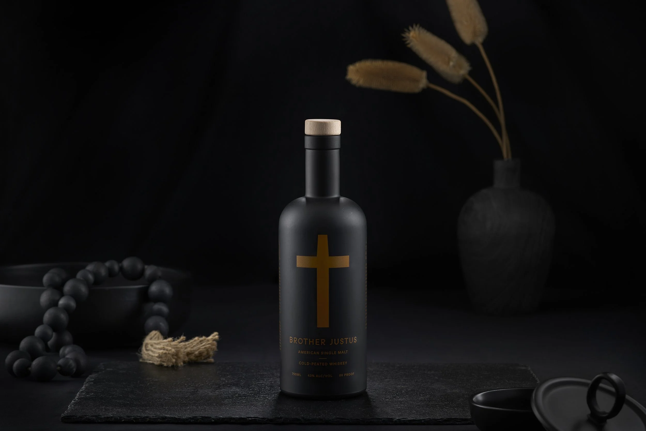 A black matte bottle of Brother Justus cold-peated whiskey with a beige cap and a gold cross on the front, placed on a dark textured surface. In the background, a vase with dried beige flowers, a black bowl with beads, and a black and gold ring are v