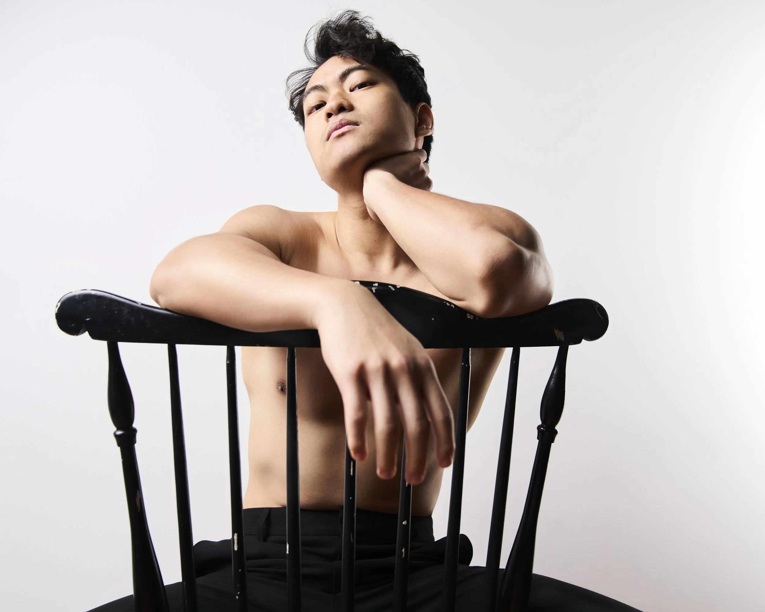 A shirtless young man with dark hair, sitting on a black wooden chair with his arm draped over the backrest, resting his chin on his hand against a plain white background.