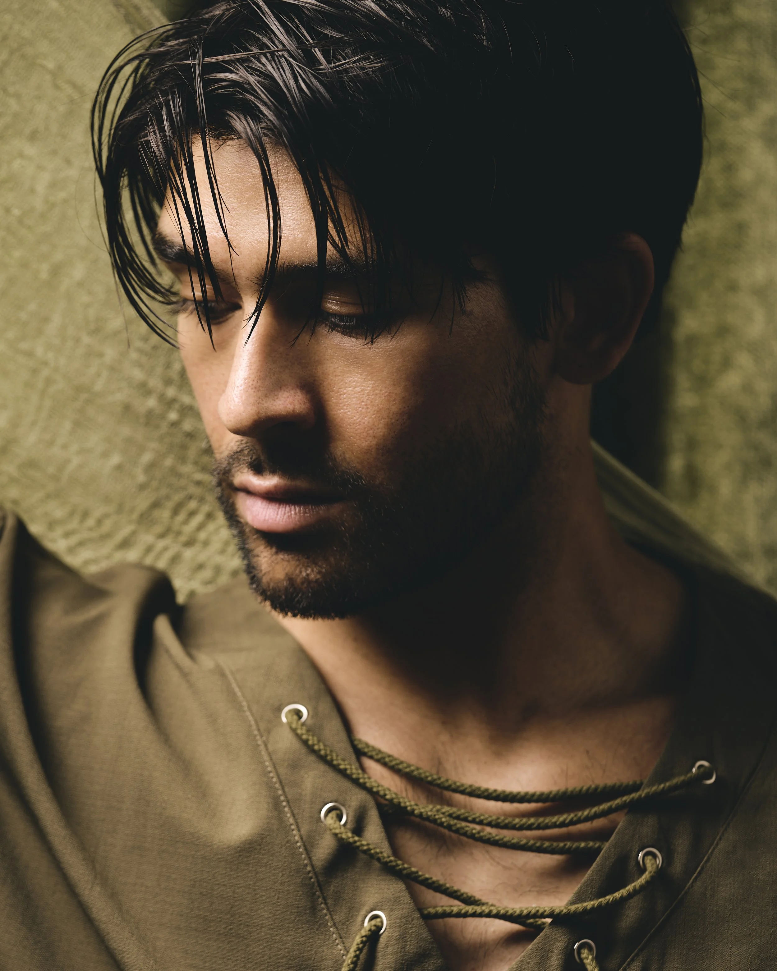 A close-up of a young man with dark, wet hair, a beard, and a neutral expression, lying against a textured olive-green background.