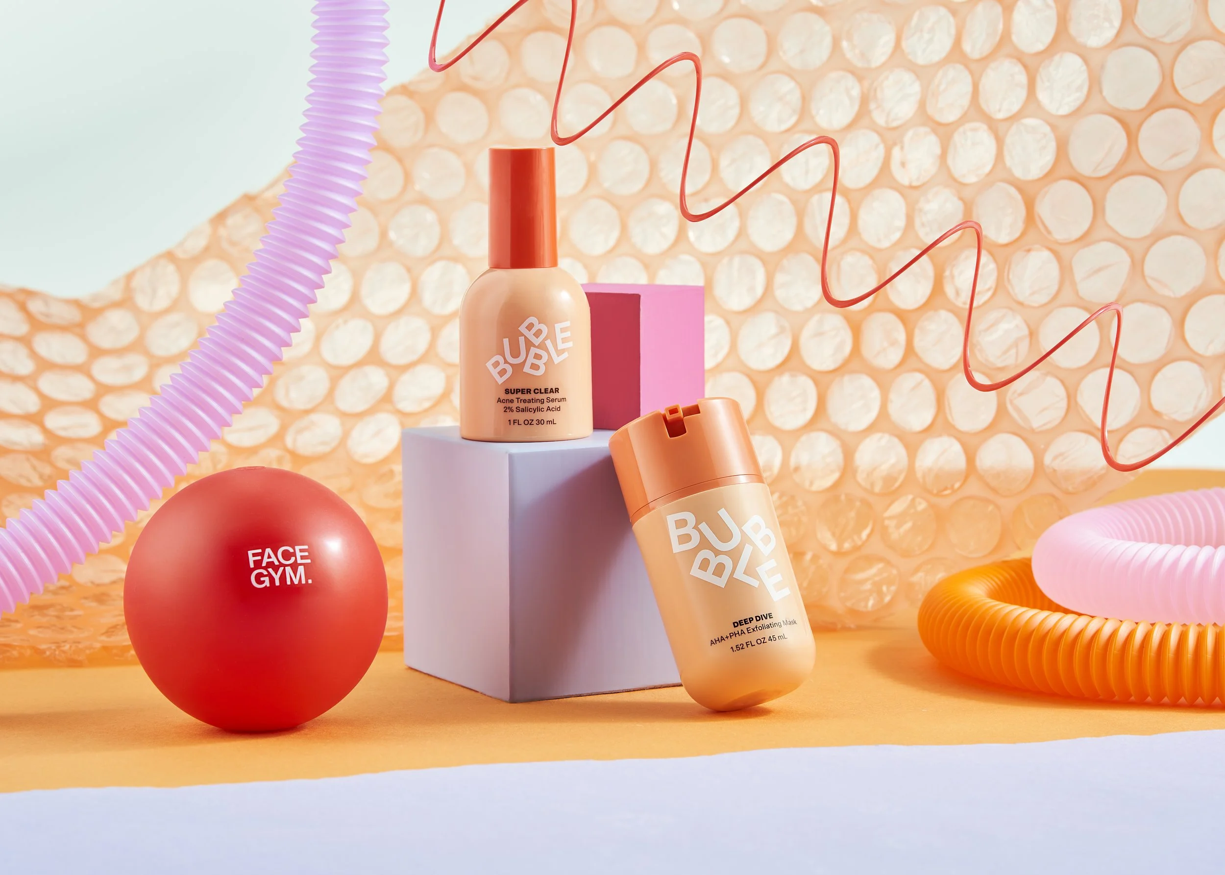 Beauty products on pastel-colored background with decorative elements including a pink sphere labeled "Face Gym," a peach serum bottle labeled "Super Clear," and a peach exfoliating mask bottle labeled "Deep Dive."