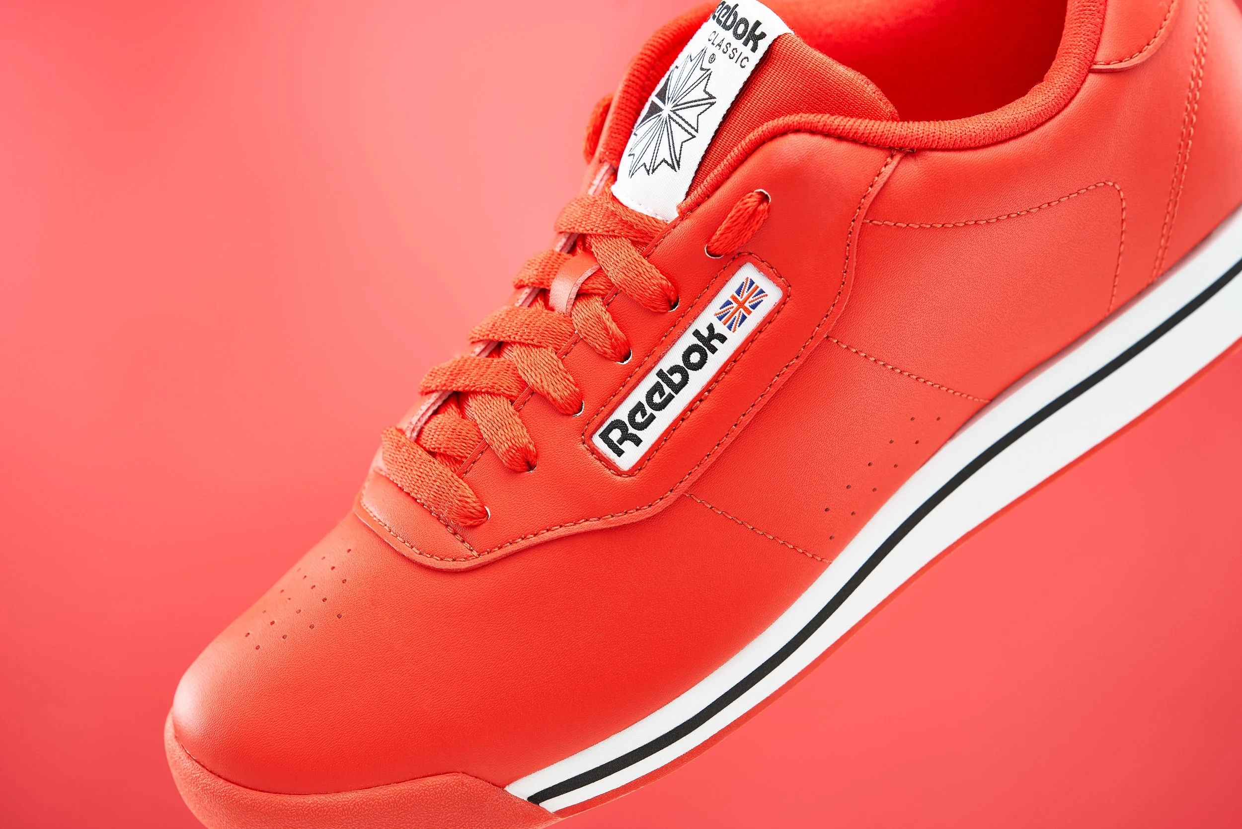 Close-up of a red Reebok Classic sneaker with white and black detailing on a pink background.