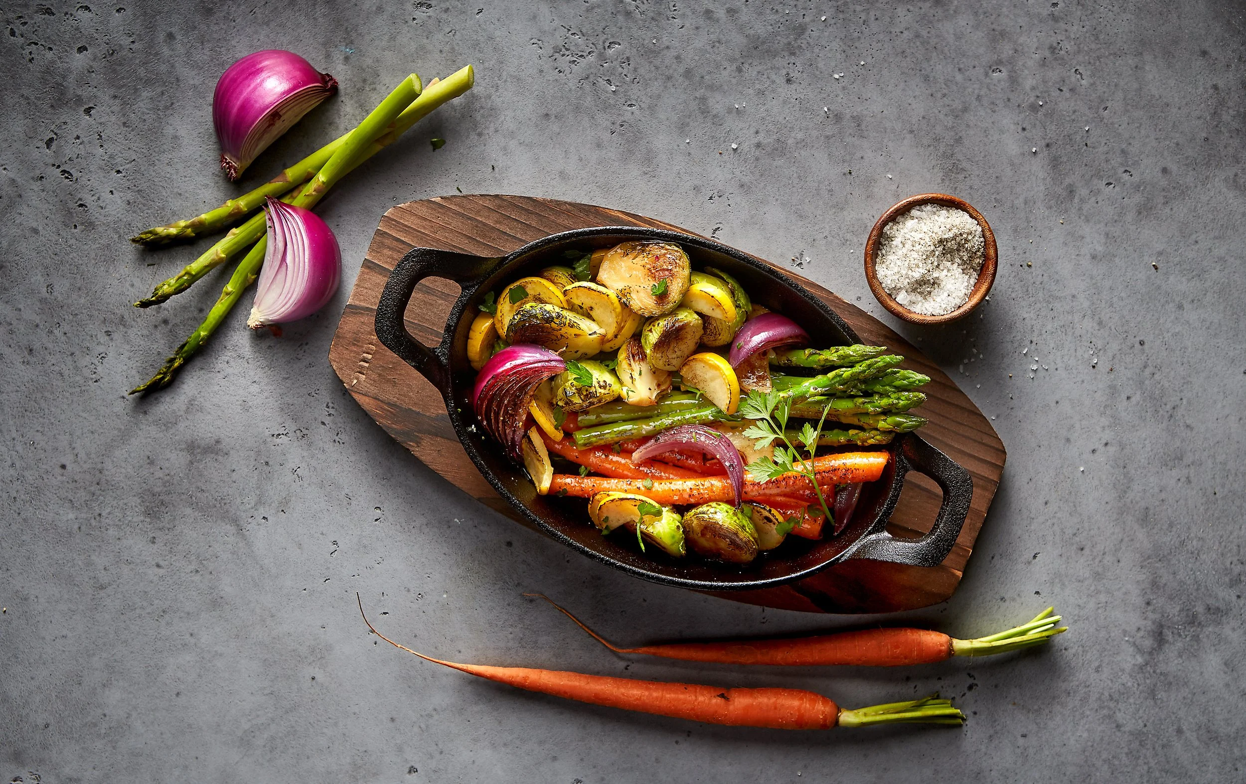 Assorted roasted vegetables including potatoes, carrots, asparagus, zucchini, and red onion in a cast iron skillet on a wooden board, with extra carrots, spring onions, and a small bowl of salt nearby on a gray surface.