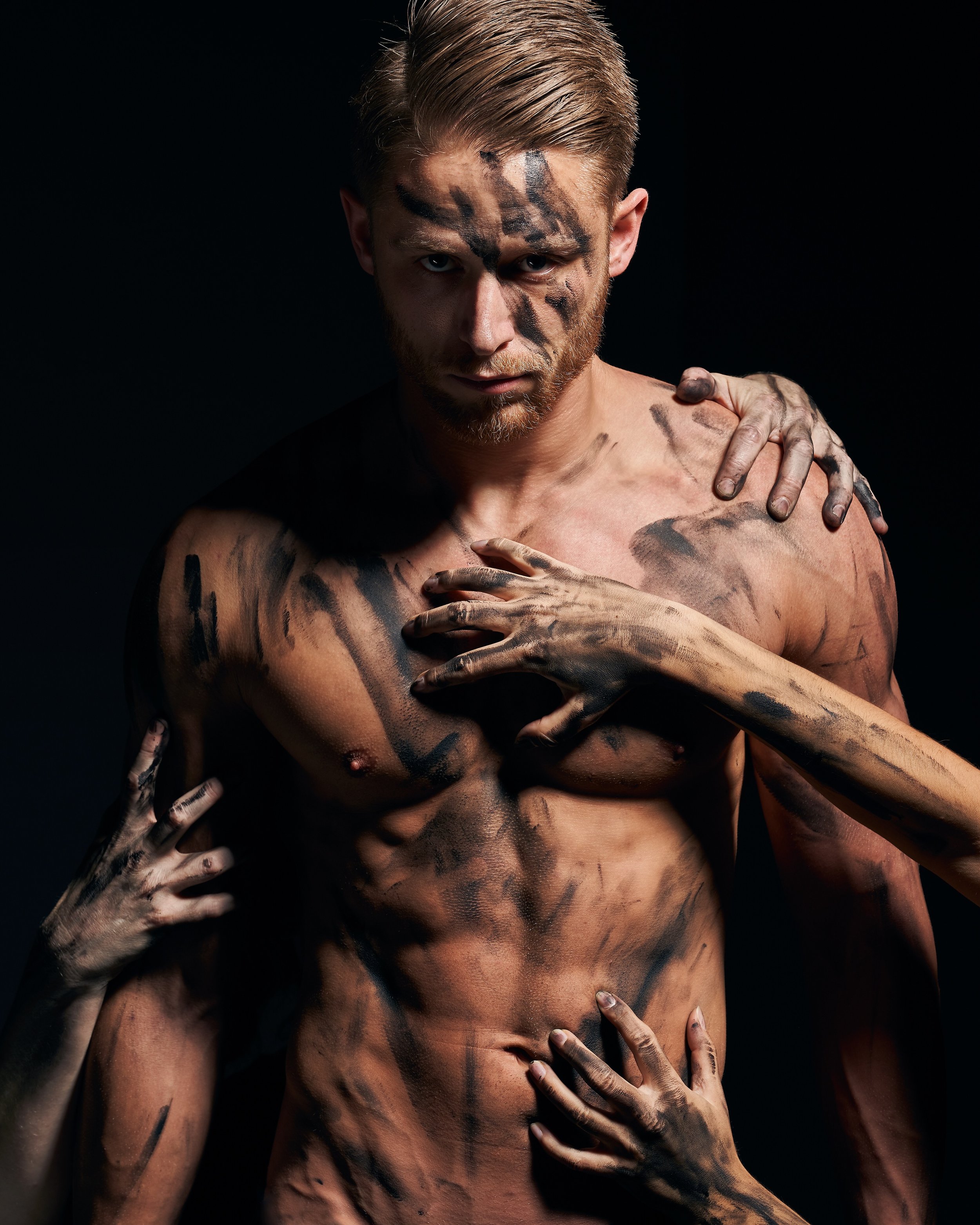 A shirtless man with light brown hair and a beard, covered in black paint strokes on his face, chest, and arms, being embraced by multiple hands from different directions against a black background.
