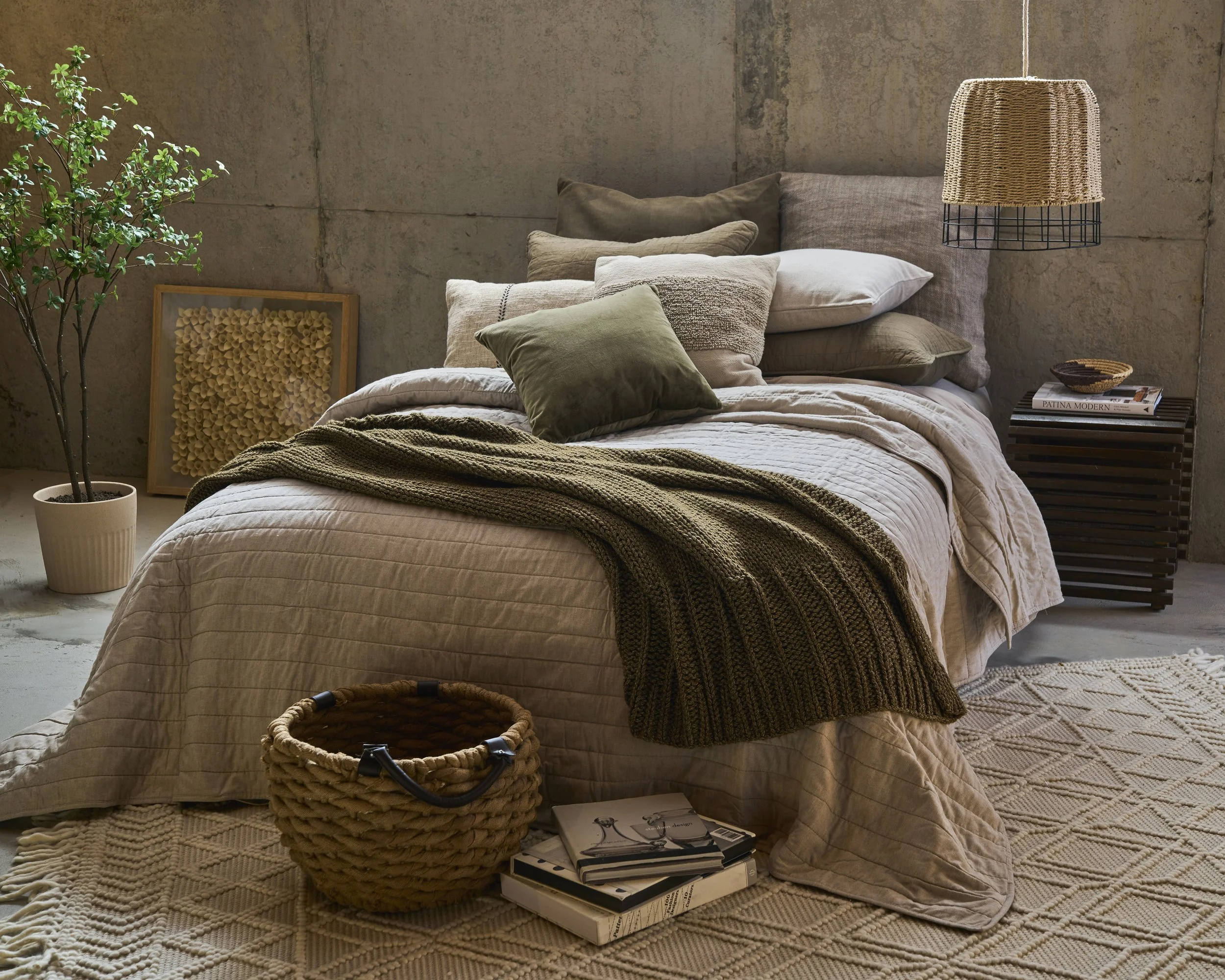A cozy bedroom with a bed covered in beige and olive green pillows and a beige quilt. A dark green knit blanket is draped over the bed. To the right, a wooden nightstand holds magazines and a small bowl, with a wicker lampshade hanging above. A potte