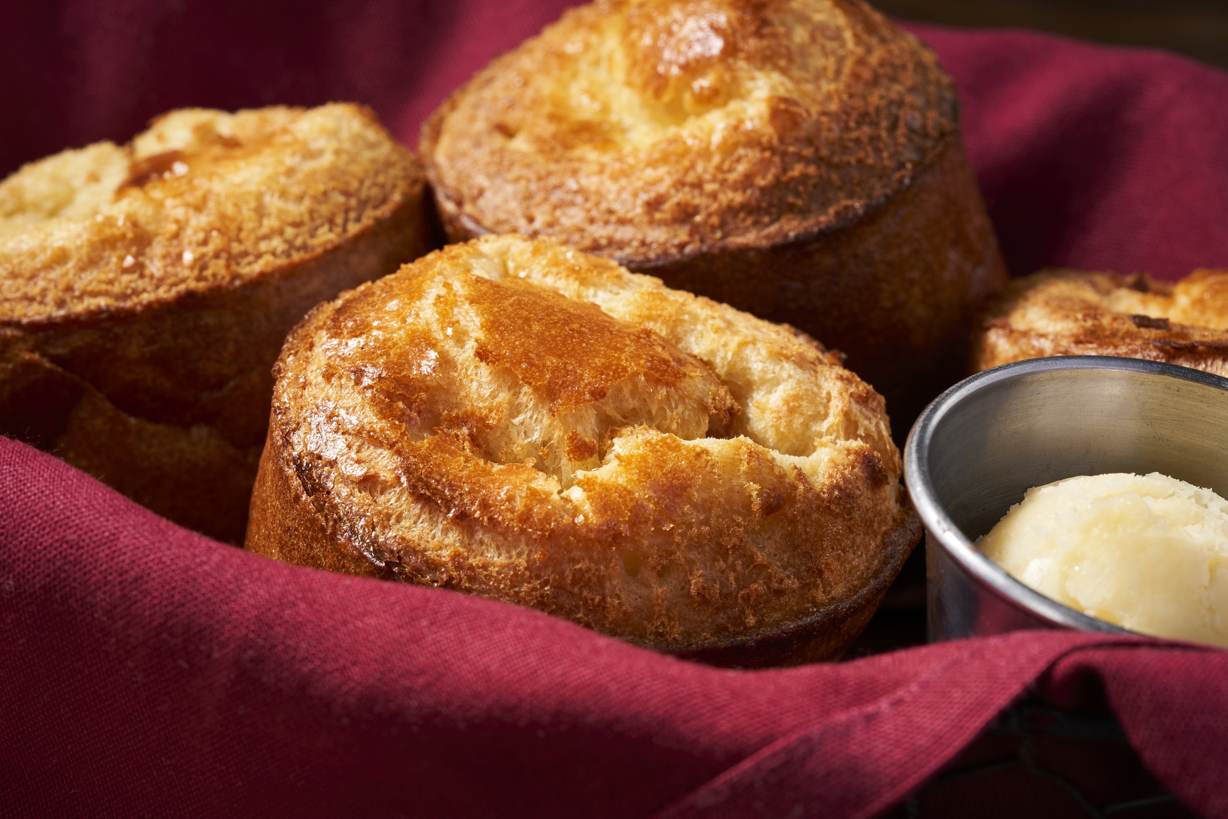 Three golden-brown muffins and a small metal cup of butter on a red cloth.