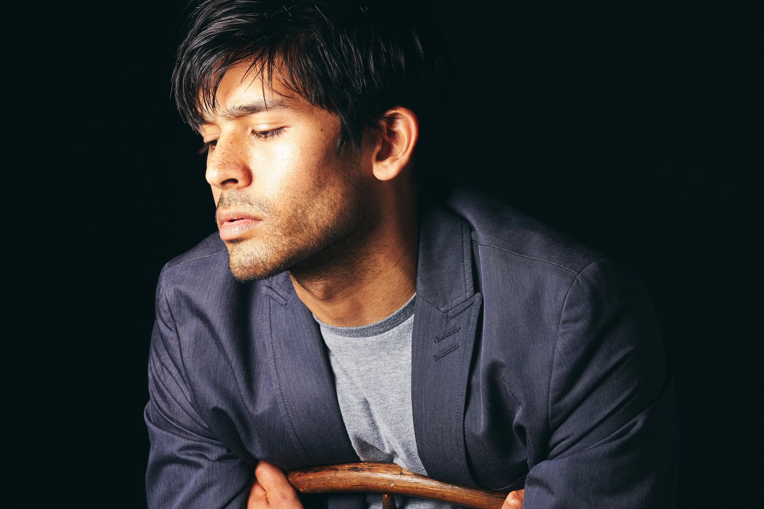 A young man with dark hair and light skin, wearing a gray shirt and a dark blazer, has his eyes closed and appears to be in deep thought against a dark background.