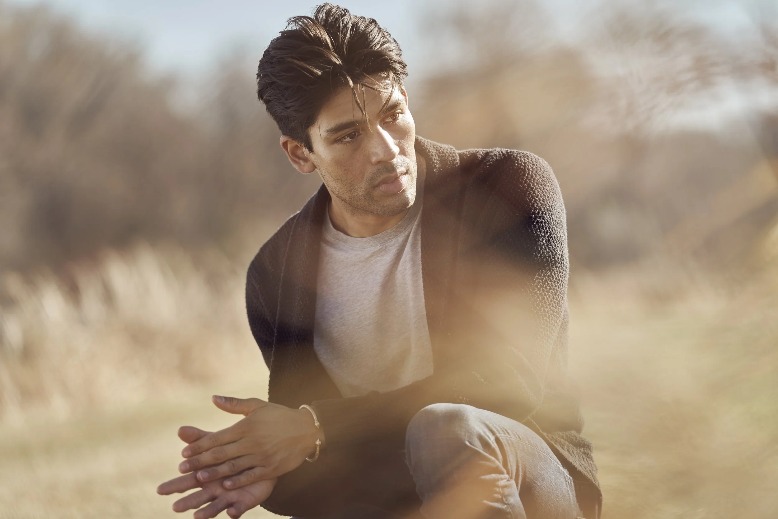 A man sitting outdoors in a natural setting with tall dry grass, looking contemplative.