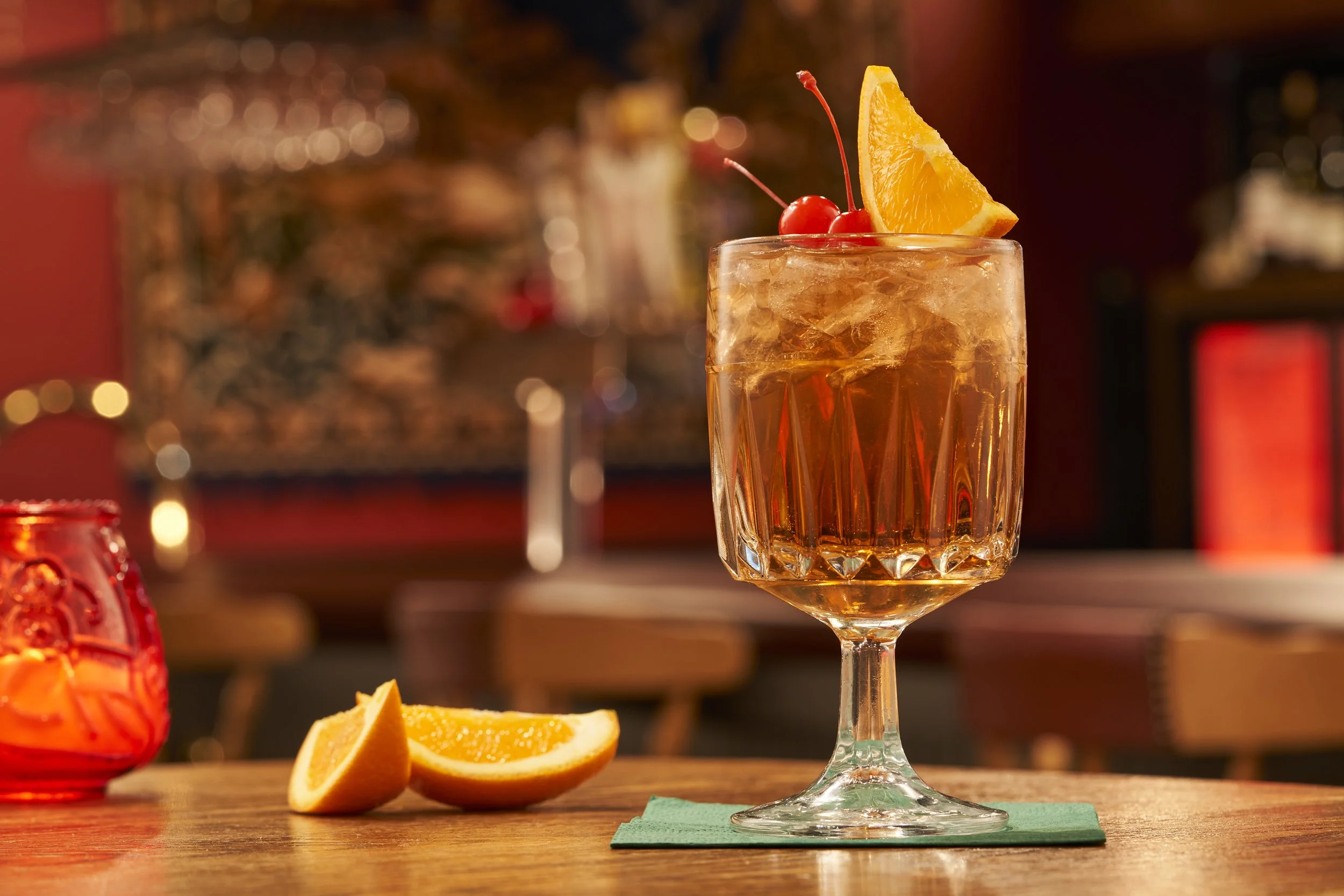 A cocktail in a glass garnished with an orange wedge and red cherries, placed on a green napkin on a wooden table with orange slices nearby, in a dimly lit bar or restaurant setting.