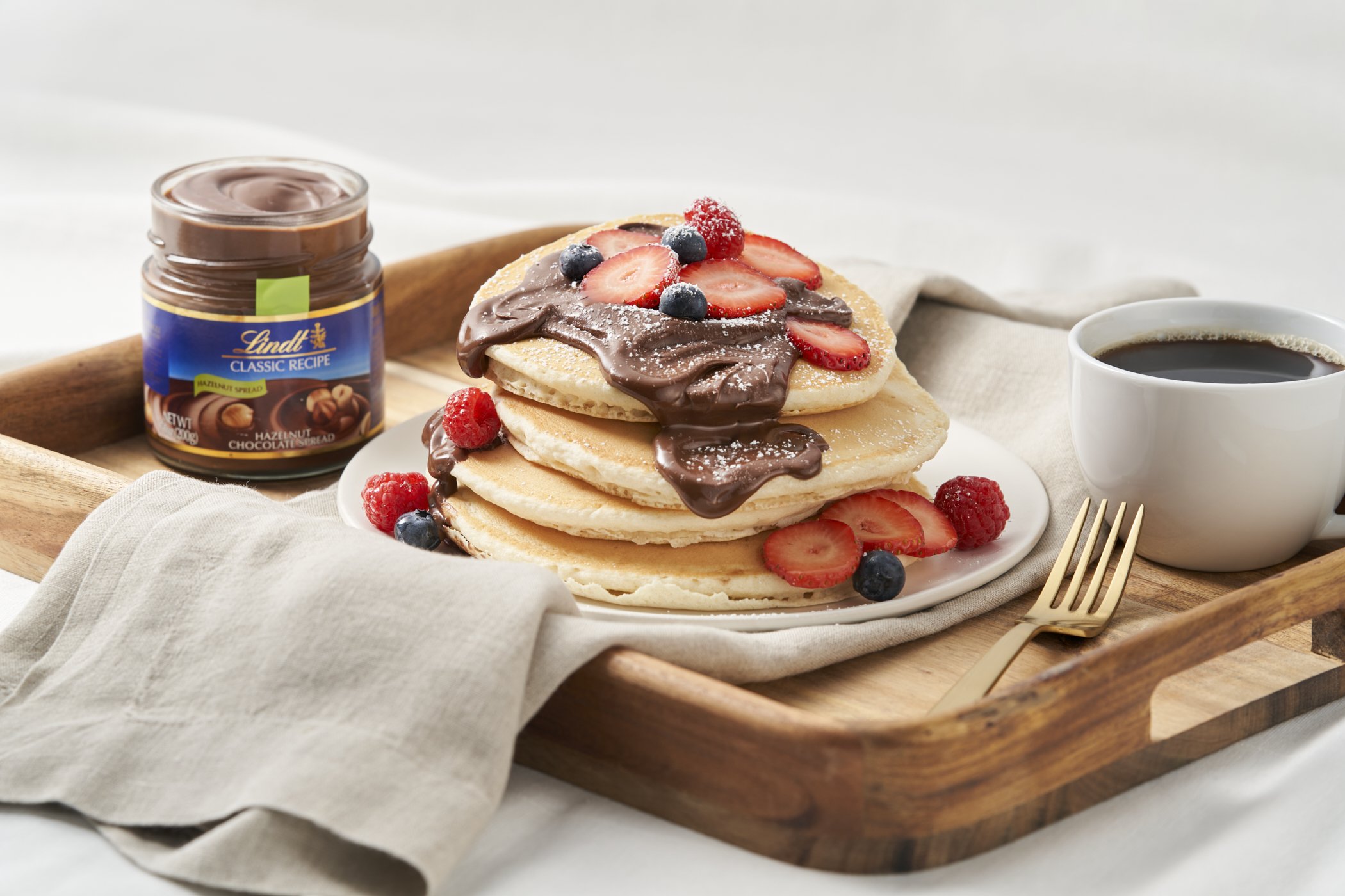 Stack of three pancakes topped with chocolate spread and fresh strawberries, blueberries, and raspberries, served with a cup of coffee and a jar of Lindt hazelnut chocolate spread on a wooden tray.