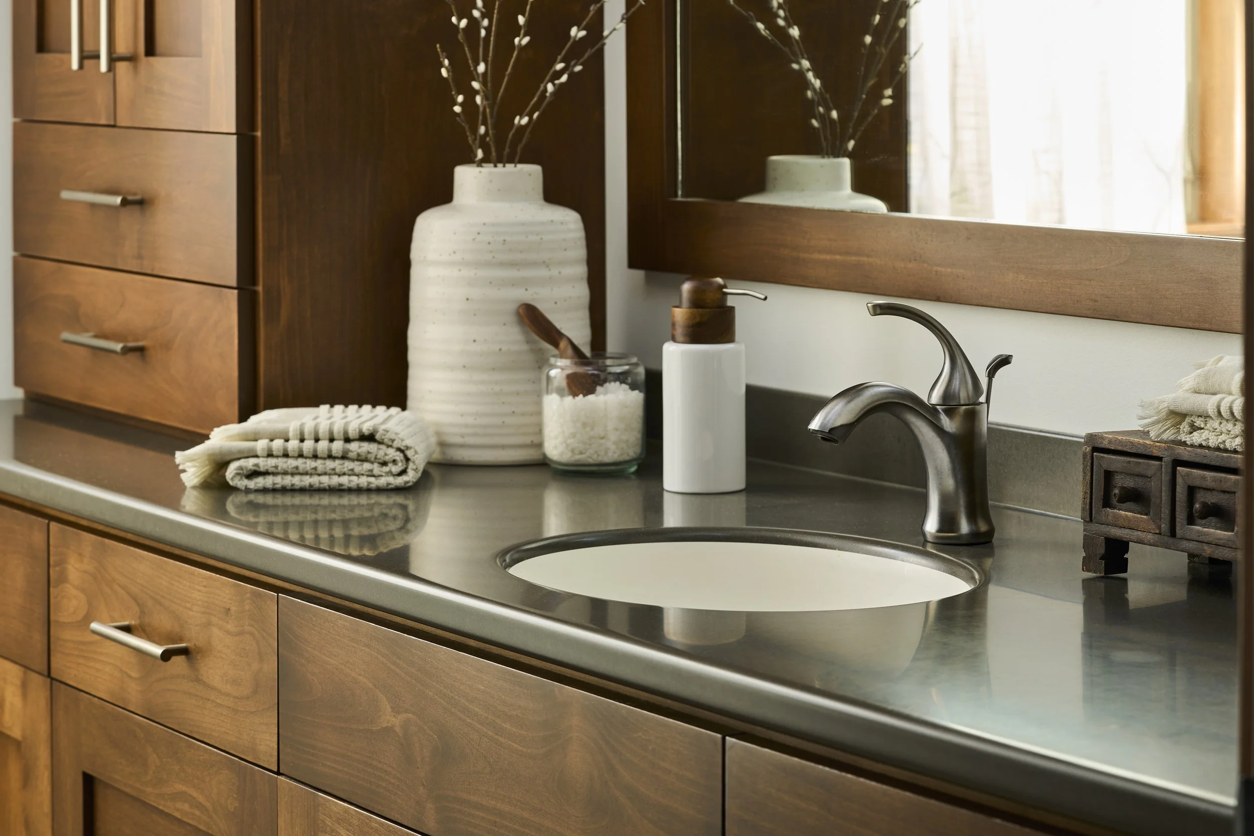 Bathroom vanity with wood cabinets, a stainless steel faucet, a large white ceramic vase with decorative branches, a soap dispenser, a jar of bath salts with a wooden scoop, and folded towels, mirrored above the sink with natural light coming through