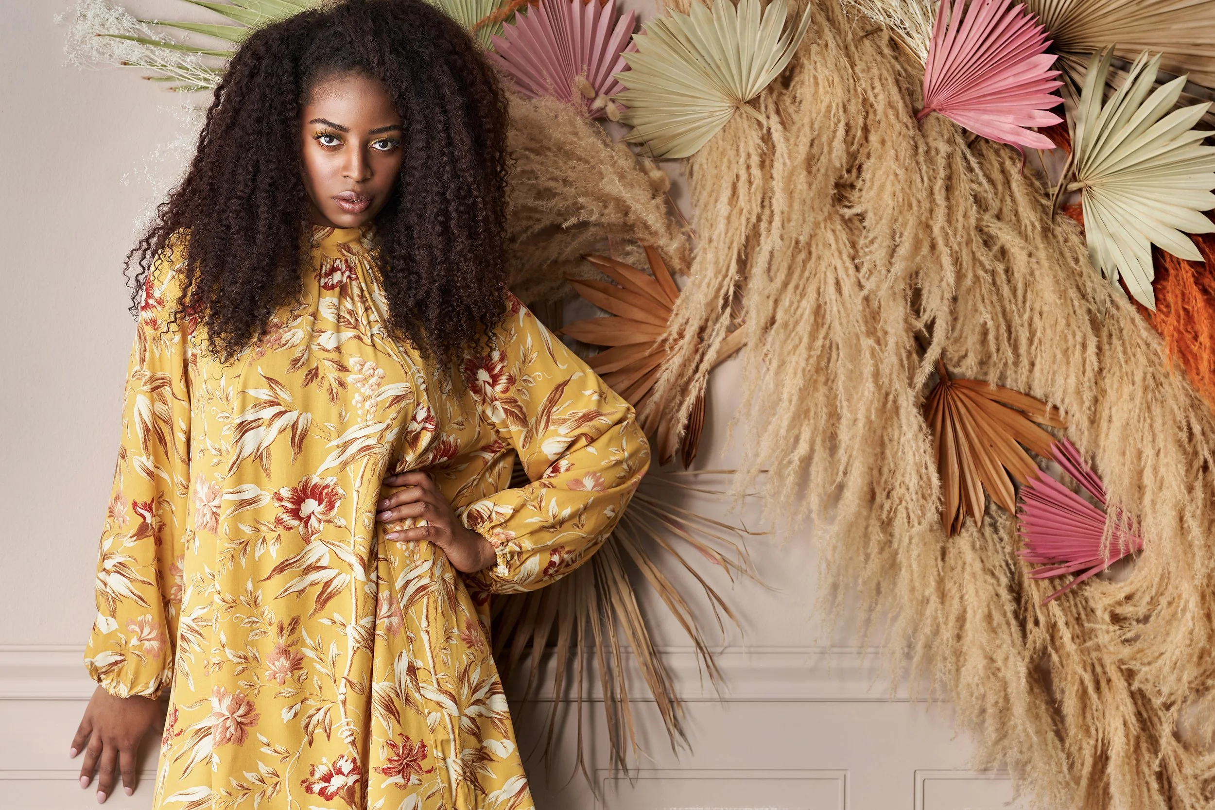 A woman with dark, curly hair wearing a yellow floral dress standing in front of a wall decorated with dried palm leaves and pampas grass in shades of pink, beige, and brown.