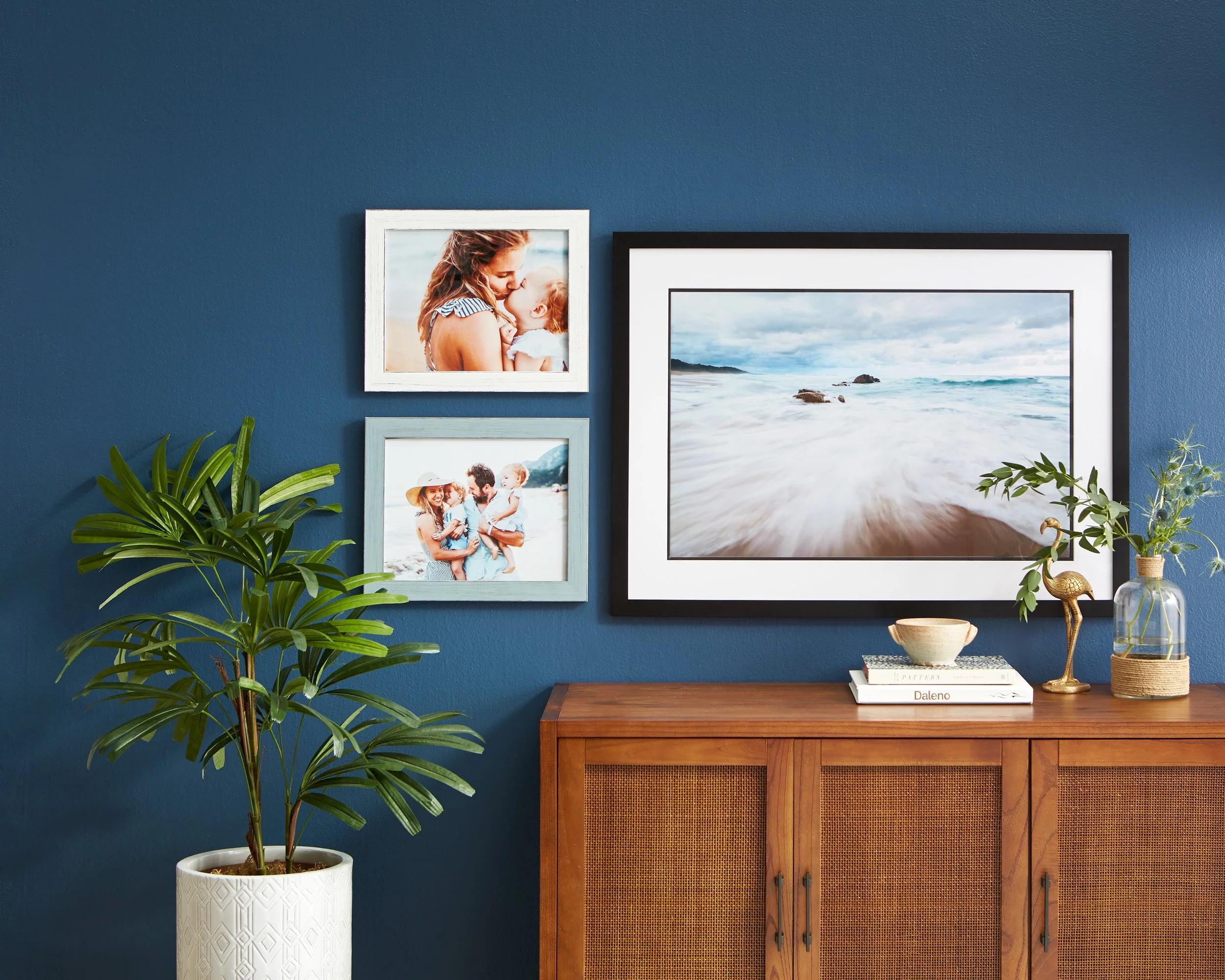 Living room with blue wall decorated with framed family and beach photographs, a potted plant, and a wooden cabinet with decorative items.