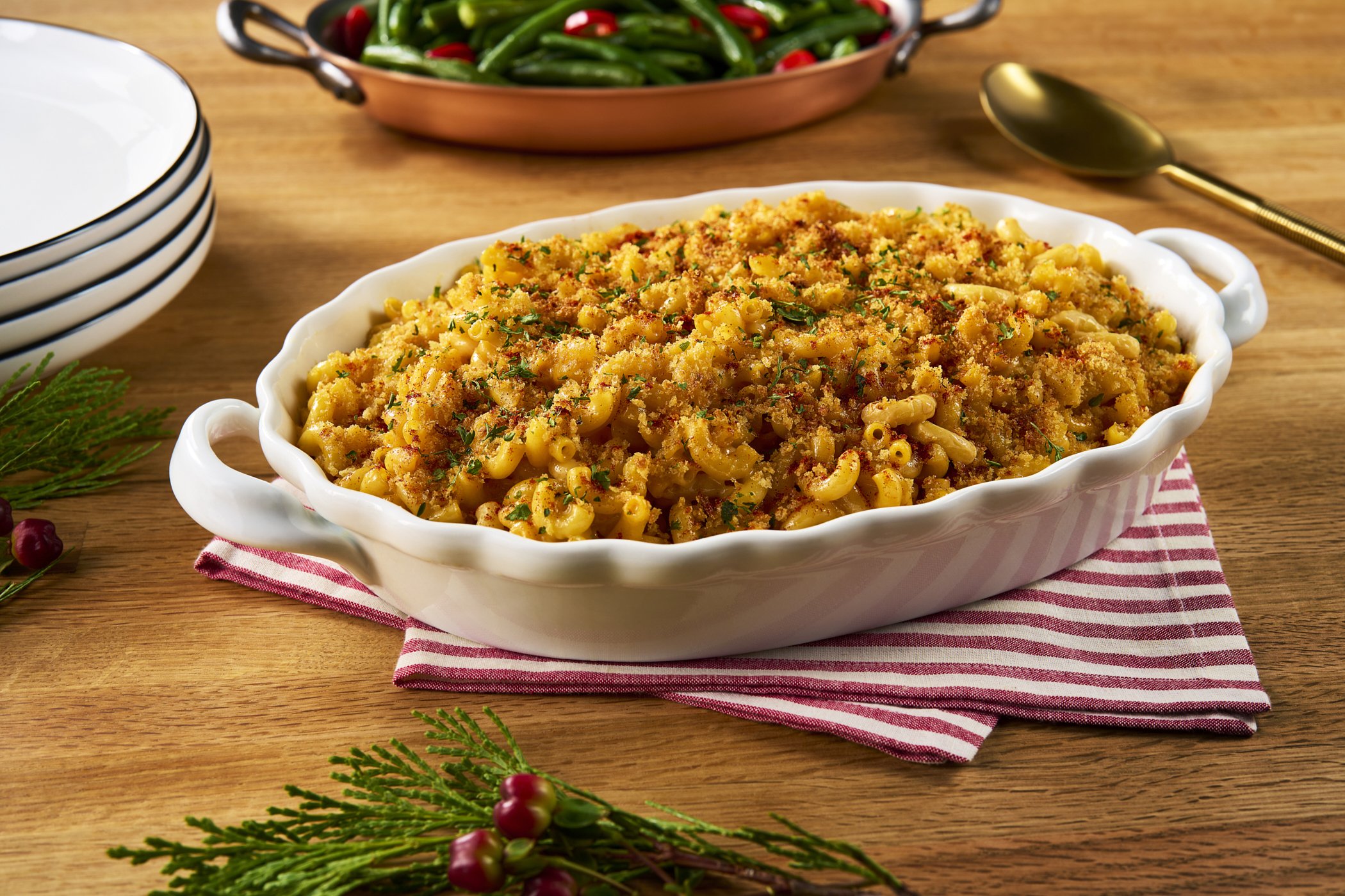 A white oval baking dish filled with macaroni and cheese topped with breadcrumbs and herbs, placed on a red and white striped cloth on a wooden table. In the background, there is a copper skillet with green beans and red chili peppers, a stack of whi
