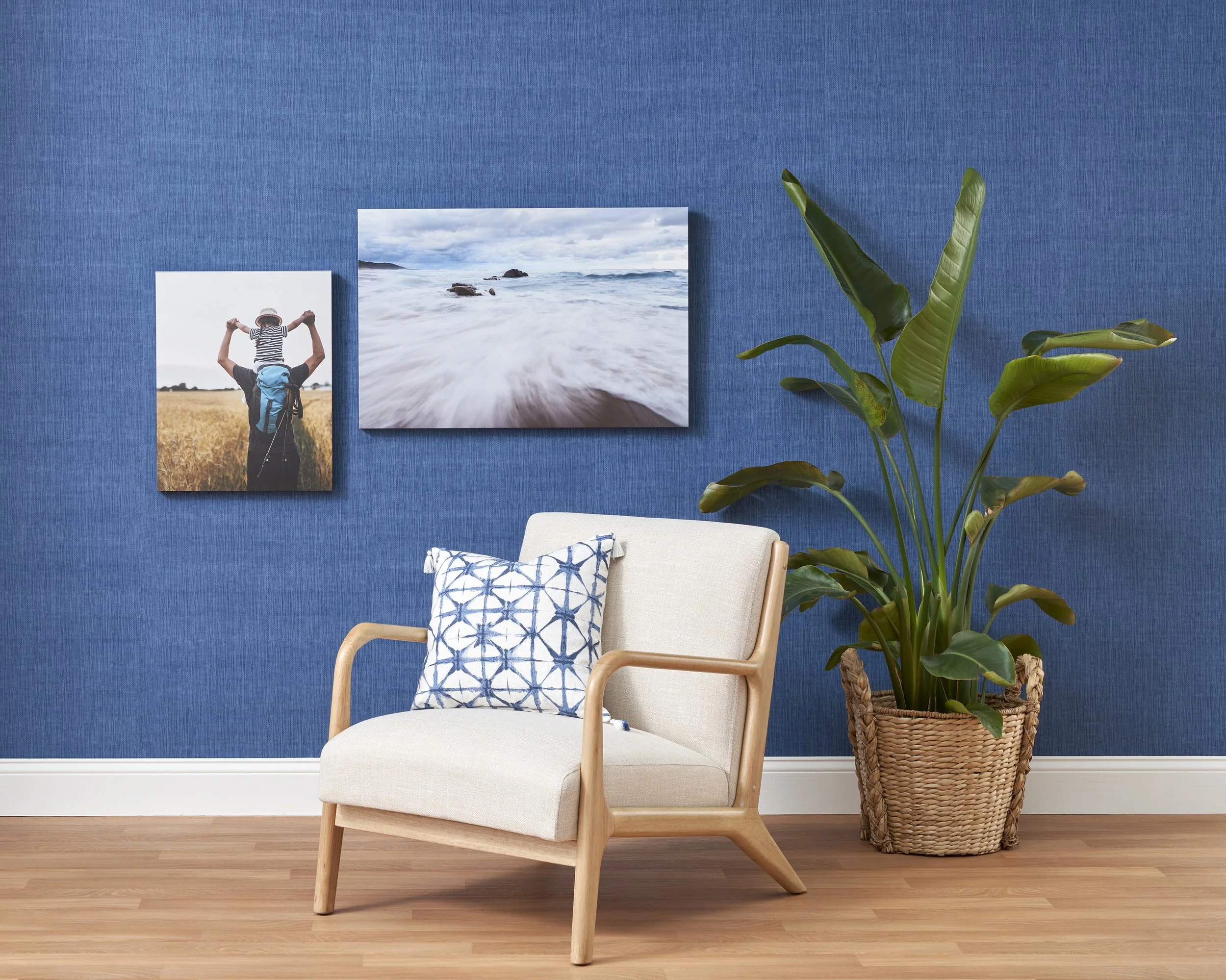 A living room corner with a beige armchair that has a blue and white patterned pillow, a large green plant in a woven basket, and two framed photos of nature on a blue wall, with a hardwood floor.