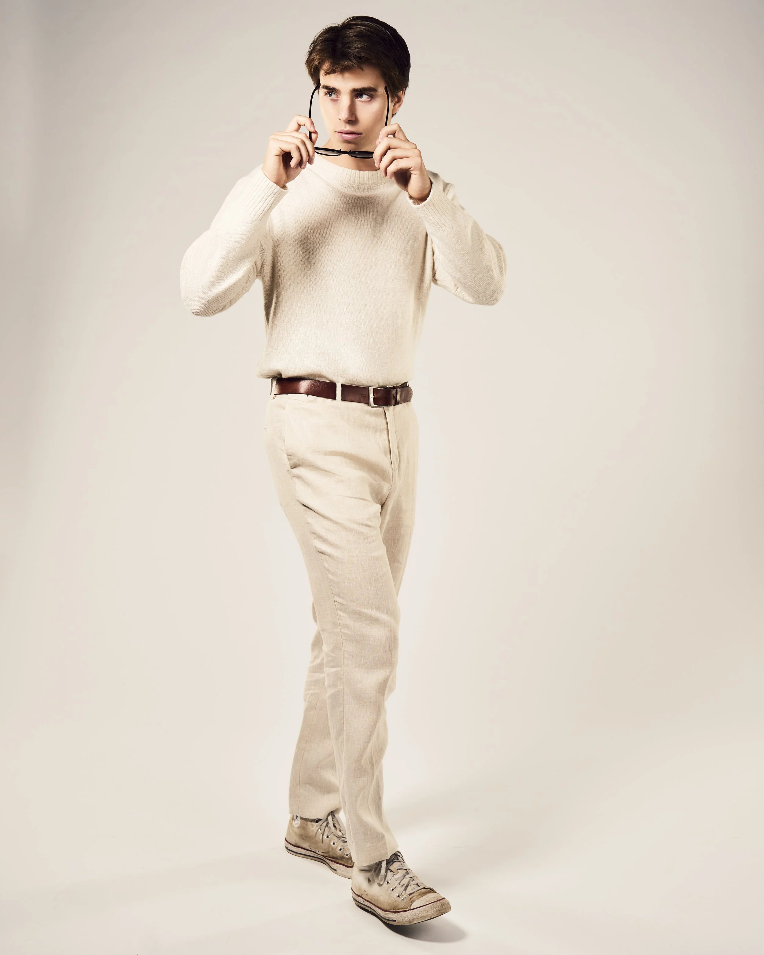 A young man with brown hair wearing a cream sweater, beige pants, gray sneakers, and holding black glasses, standing against a plain light-colored background.