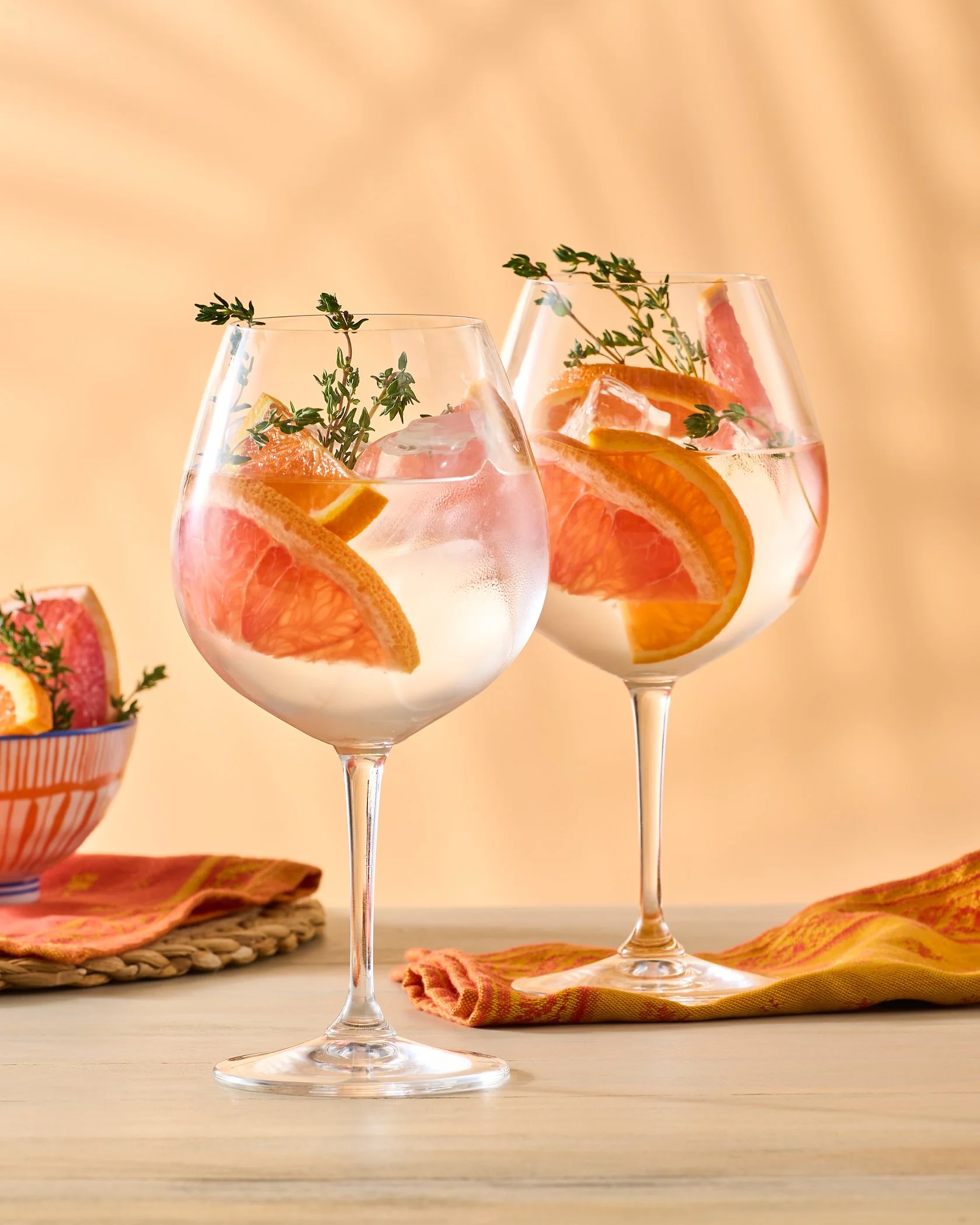 Two glasses of grapefruit spritzers garnished with sprigs of thyme on a wooden table with colorful napkins and a bowl of grapefruit slices in the background.