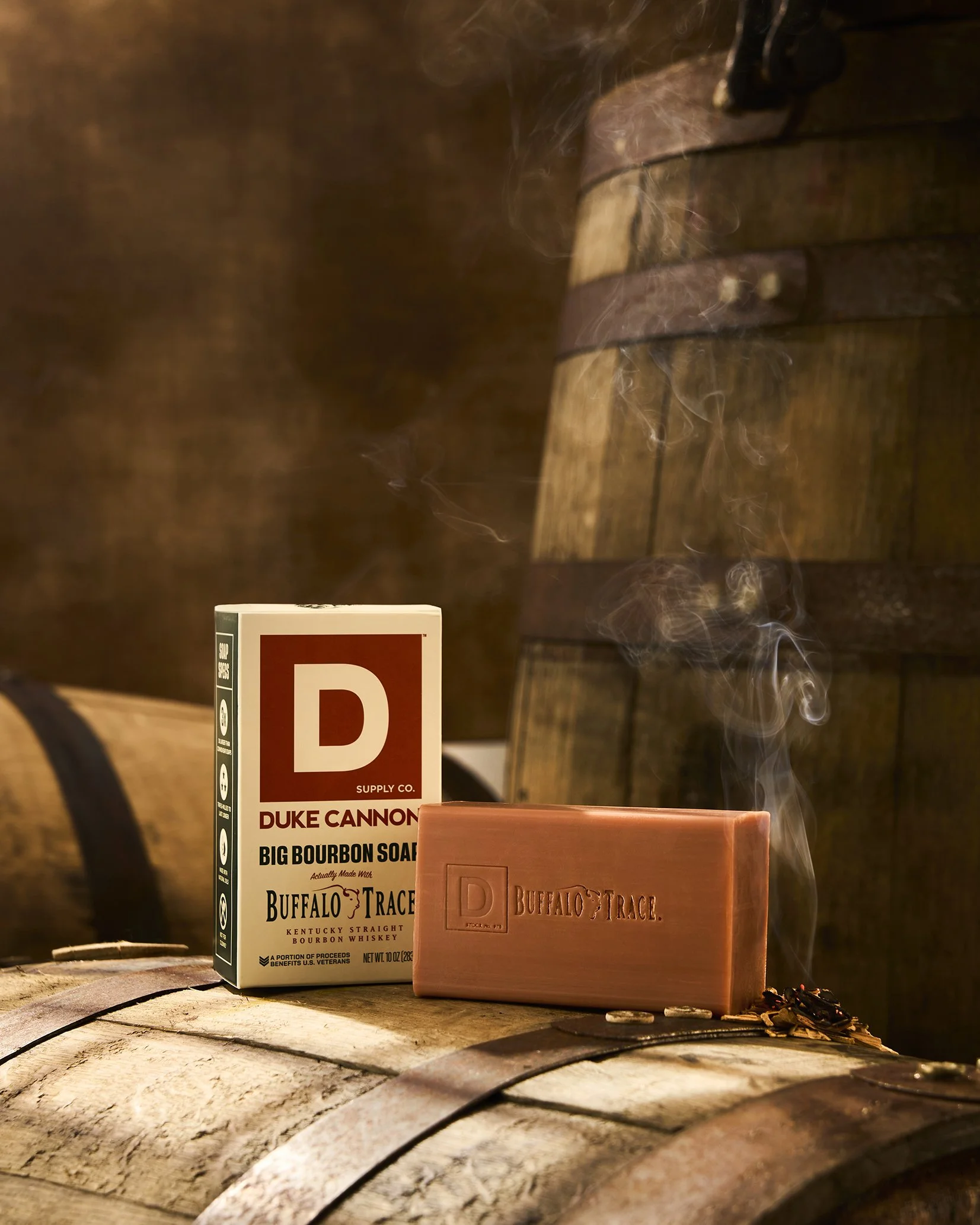 A bar of Buffalo Trace bourbon soap with steam rising next to a box of Duke Cannon Big Bourbon Soap, placed on a wooden barrel in a rustic setting.