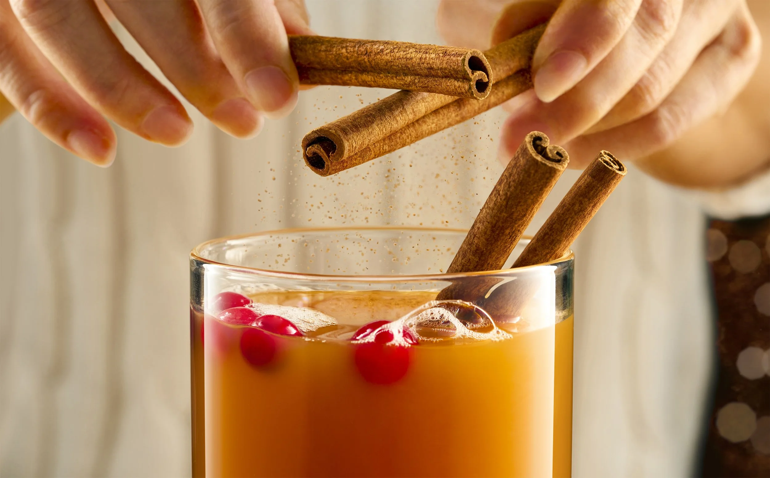 Hands dropping cinnamon sticks into a glass of orange-colored beverage, with red berries and ice cubes, possibly a mulled drink or holiday punch.