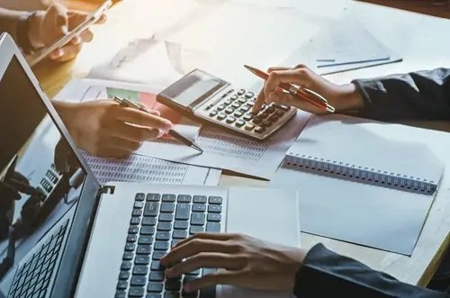 Two people working at a desk with laptops, calculators, papers, and notebooks, discussing business or finance.