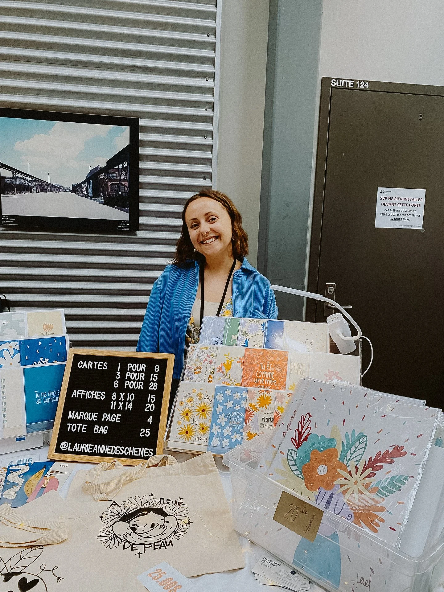 Une femme souriante se tient derrière un stand de vente de cartes, affiches et tote bags avec des illustrations florales et des messages de soutien, dans un environnement intérieur.