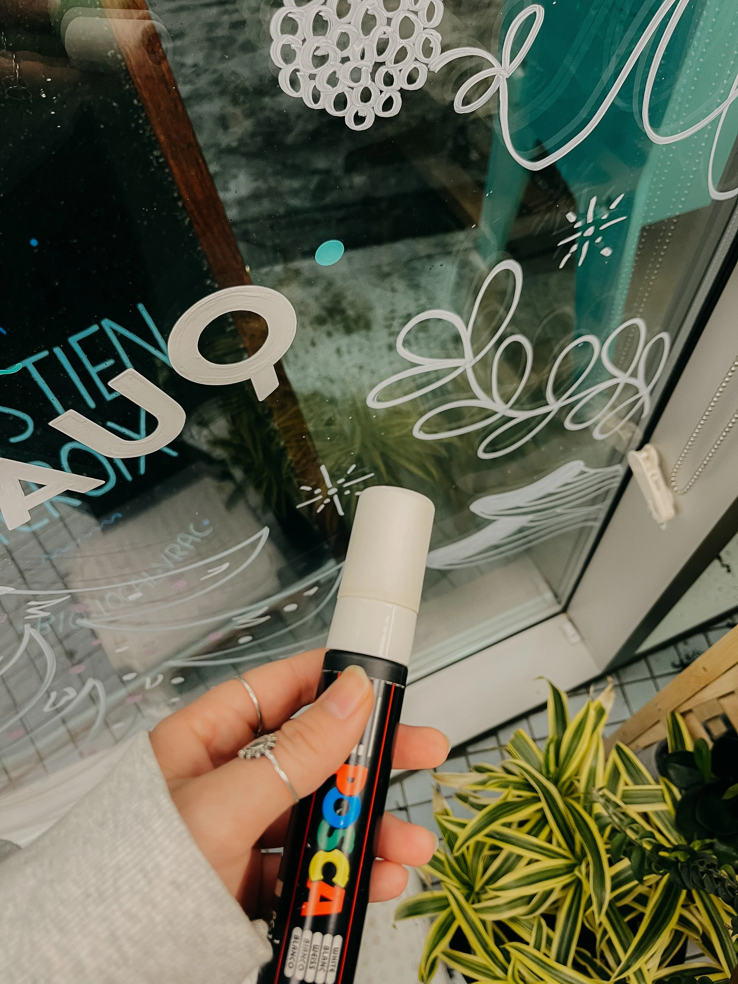 Mains tenant un marqueur blanc à tip en mousse, dessinant des décorations florales et un texte en français sur une vitre, avec des plantes en pot en dessous.