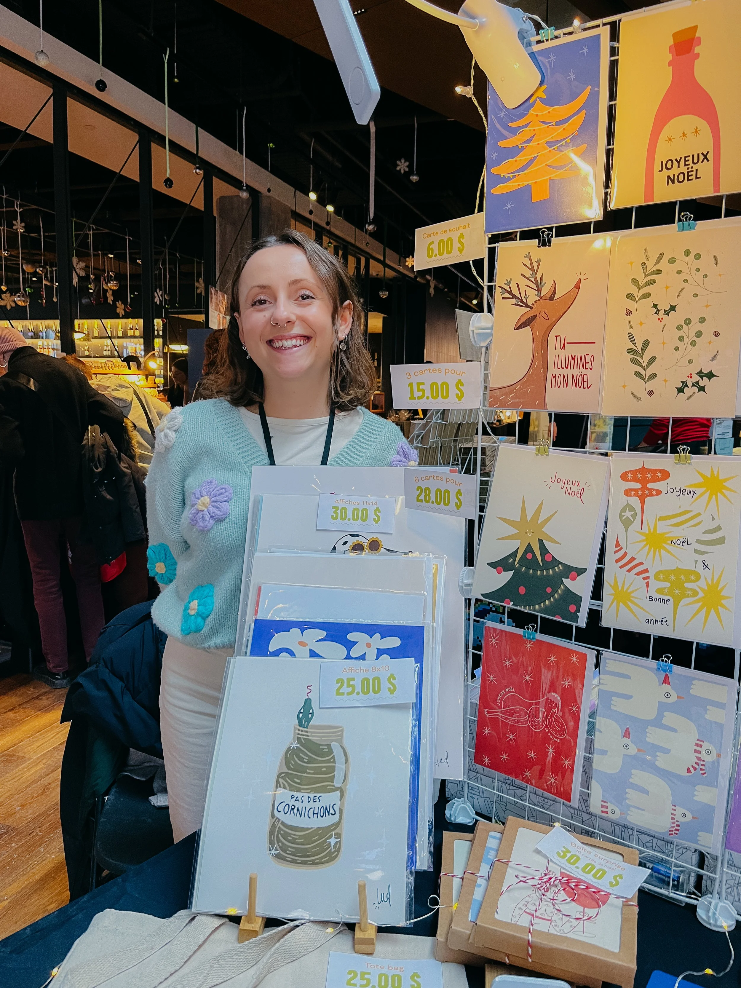 Une femme souriante devant un stand de cartes de vœux de Noël décoré avec des affiches aux motifs festifs et des prix.