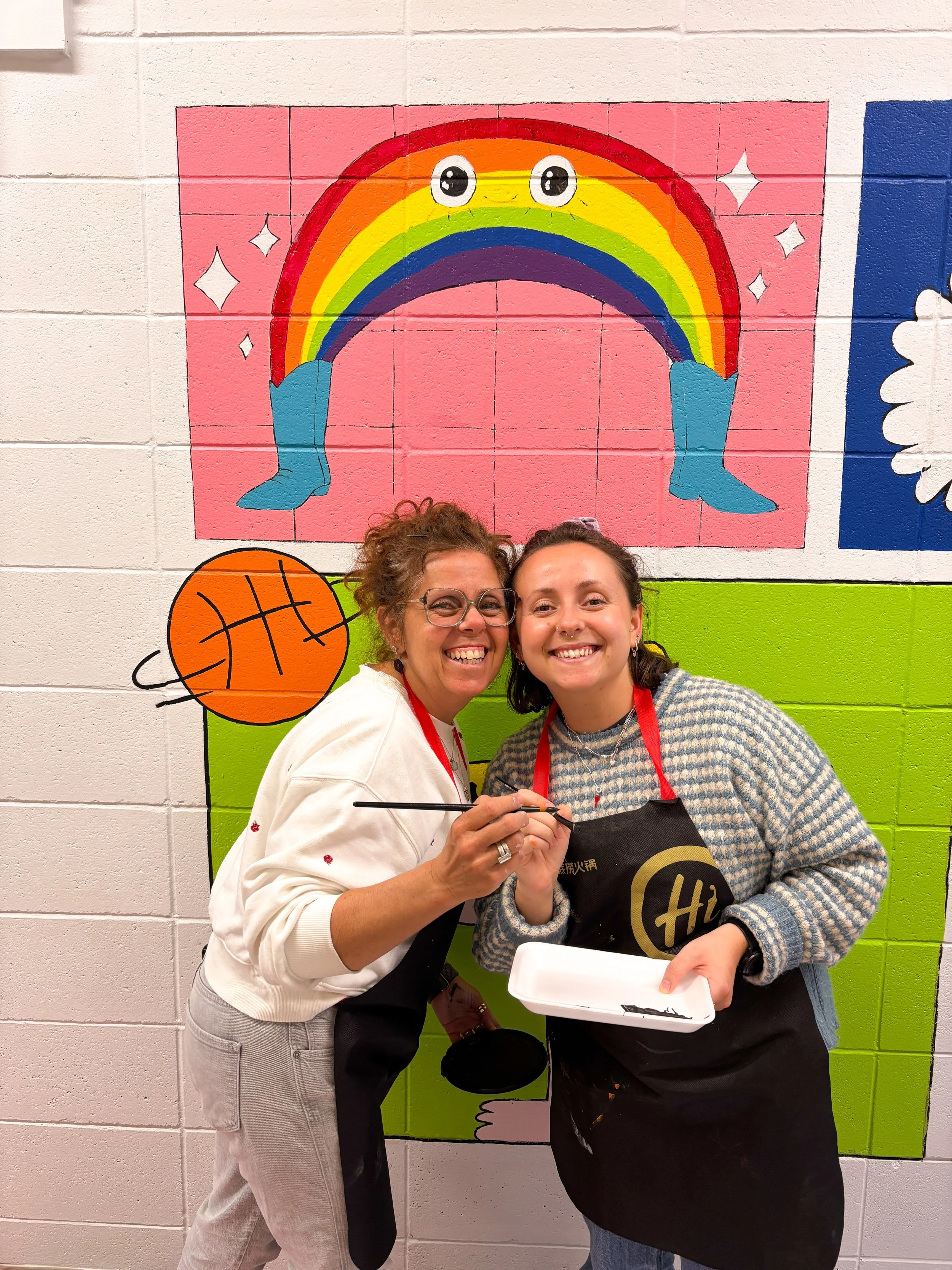 Deux femmes souriantes posent devant un mur peint avec un arc-en-ciel aux yeux yeux emoji et un ballon de basket. Elles portent des tabliers et semblent participer à une activité artistique.