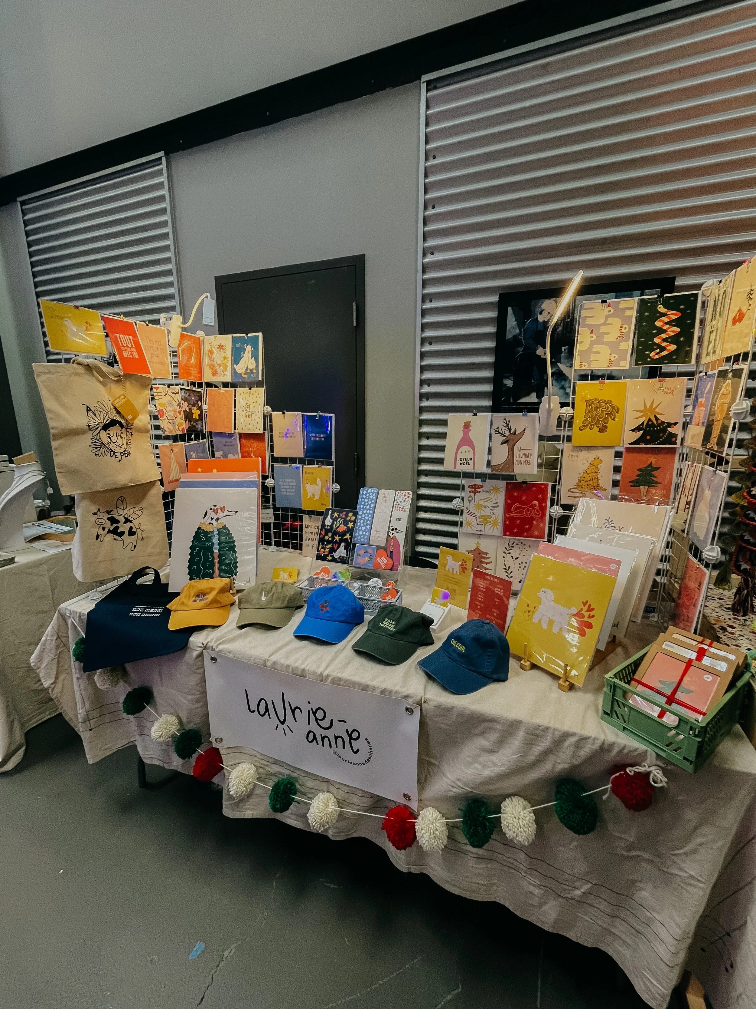 Stand de vente d'articles de Noël, avec des cartes, des casquettes, un sac en toile, et une guirlande de pompons, à l’intérieur d’un bâtiment avec un fond de panneaux en métal.