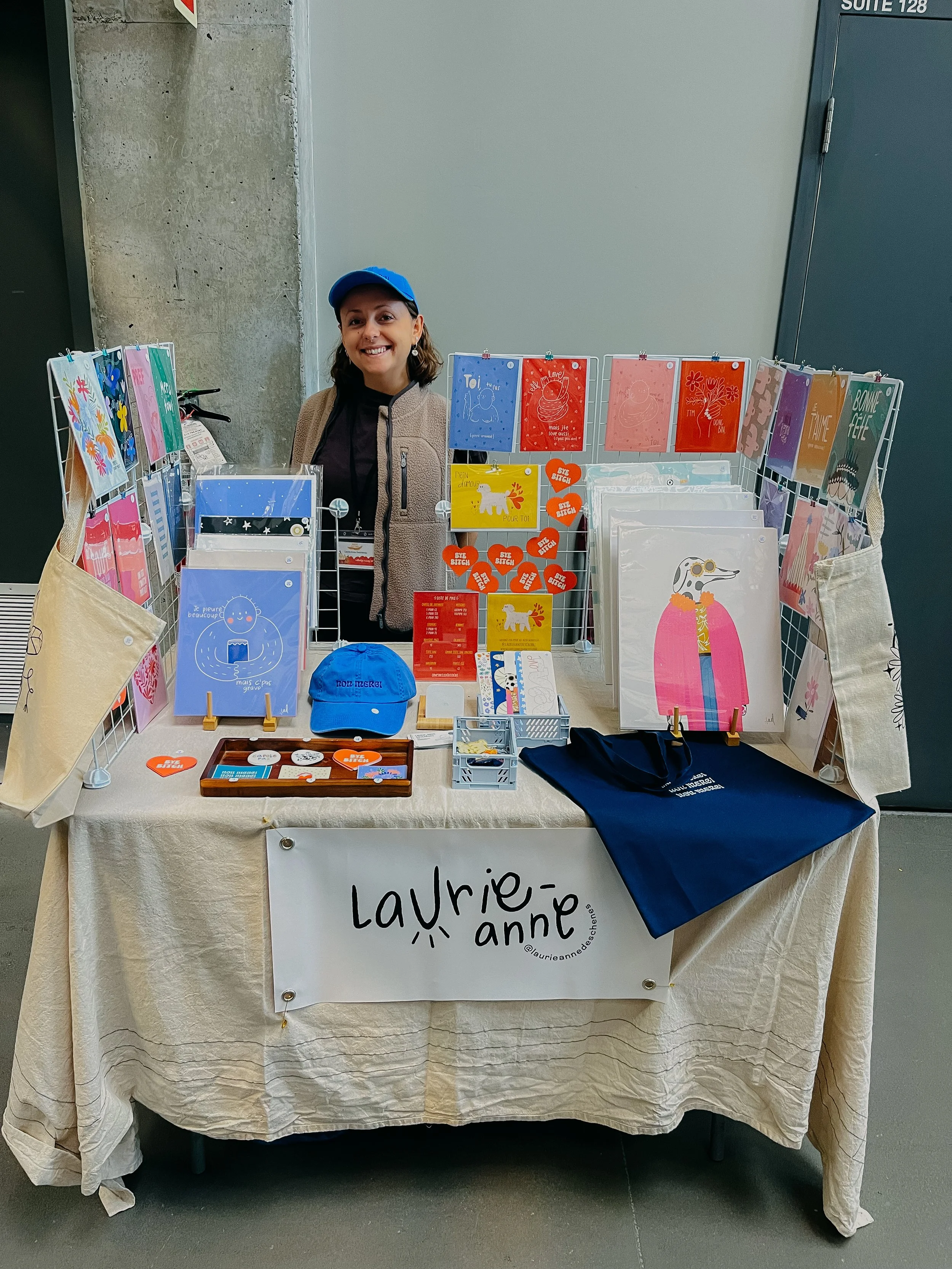 Une femme souriante derrière un stand de vente d'articles colorés, comme des cartes, des affiches, un chapeau bleu, une sacoche, et des objets décoratifs, avec un panneau portant le nom 'Laurie-anne'.