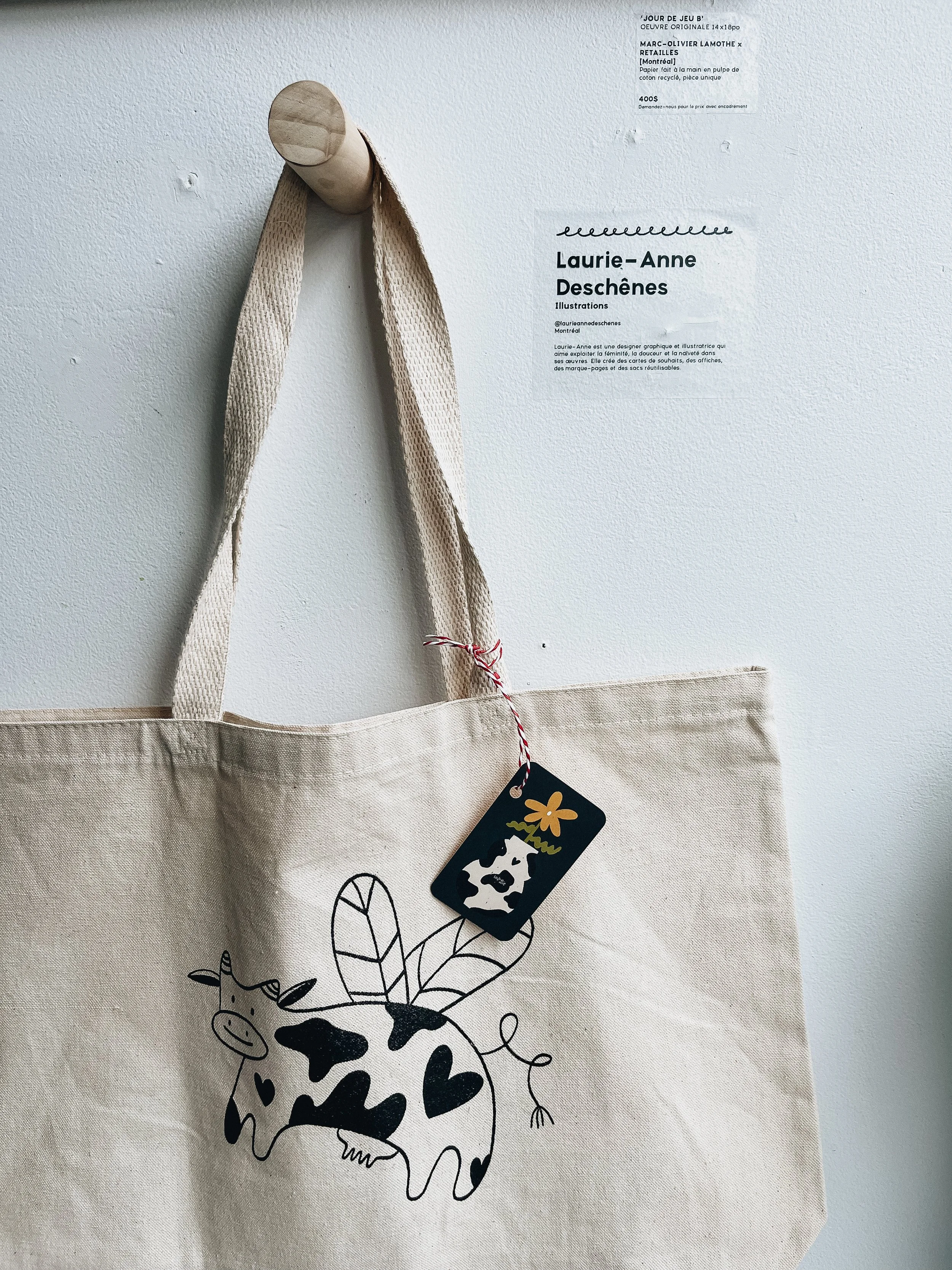 Tote bag en toile avec illustration d'une vache stylisée, décorée de cœurs noirs, avec des feuilles vertes et une étiquette en plastique noir attachée à la poignée.