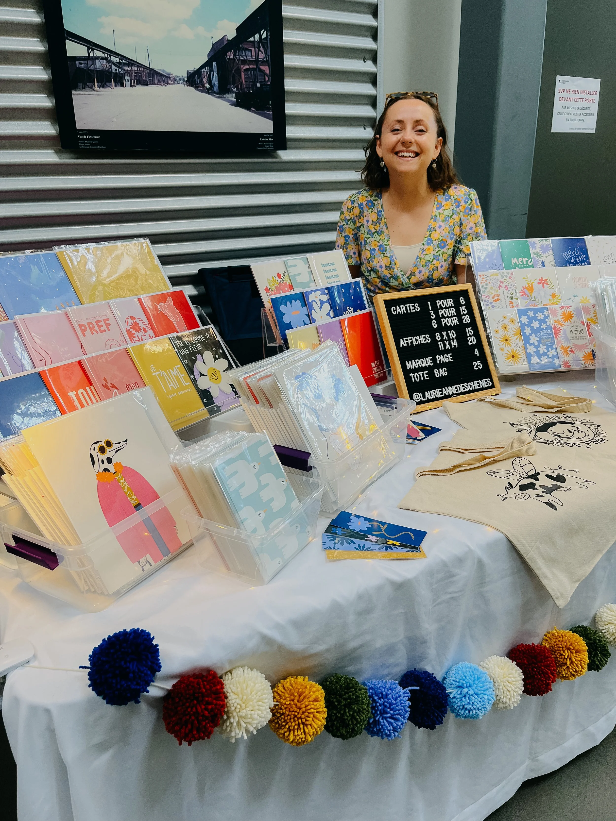Une jeune femme souriante derrière un stand de vente de cartes, affiches et tote bags, décoré avec des pompons colorés à la bordure d'une table.